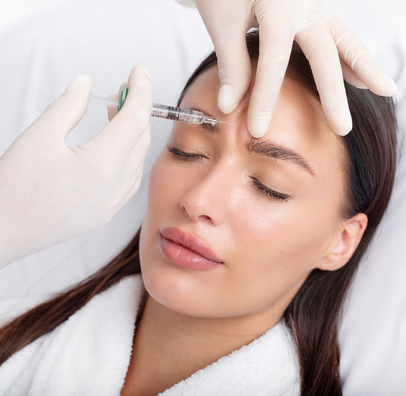 Woman receiving a cosmetic injection between eyebrows, lying down with eyes closed. Gloves and syringe visible.