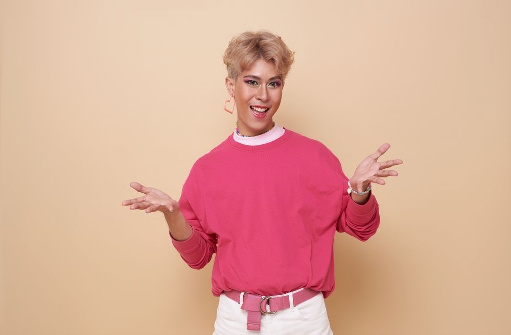 Person with short blonde hair and pink clothing gesturing with hands, smiling against a beige background.