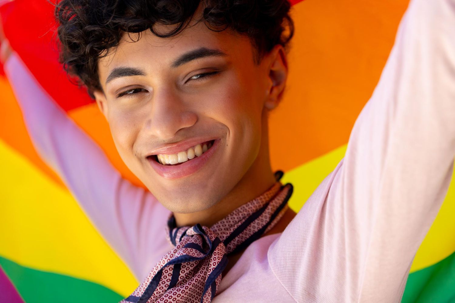 A young man is holding a rainbow flag in his hands and smiling.