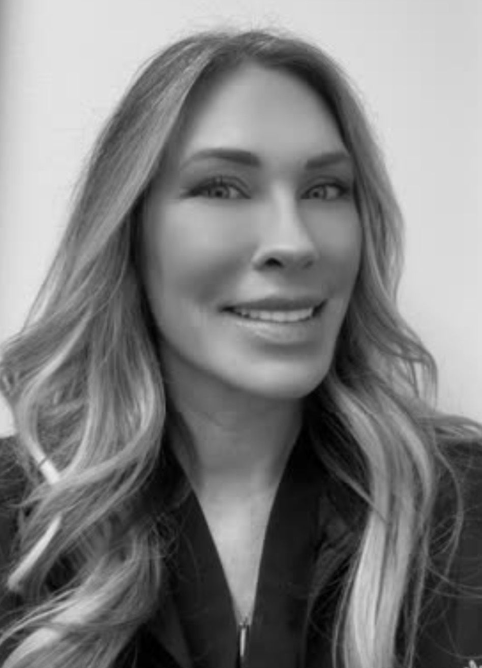 Woman with long, wavy blonde hair smiling, wearing a dark top, in a well-lit space.