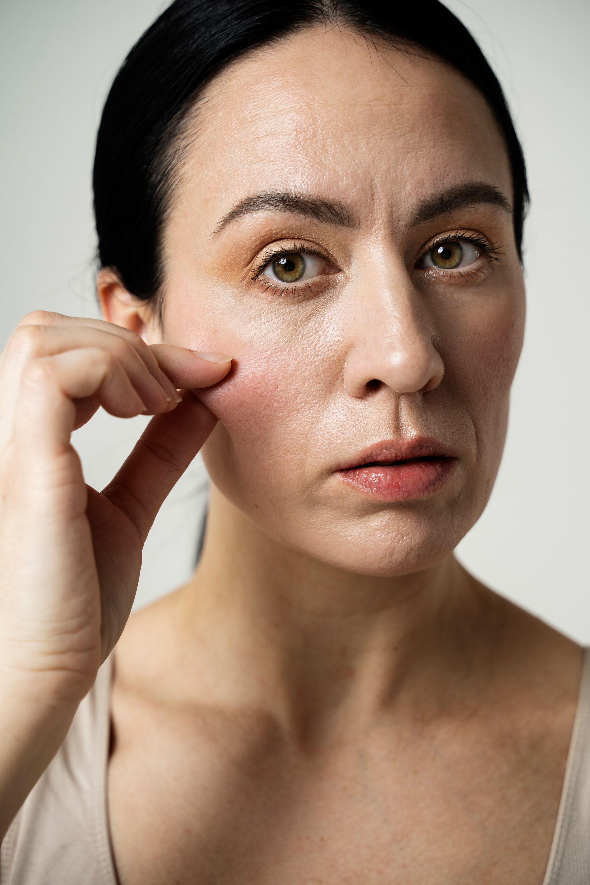 Woman pinching her cheek, looking at the camera, with visible skin texture and wrinkles, against a white background.