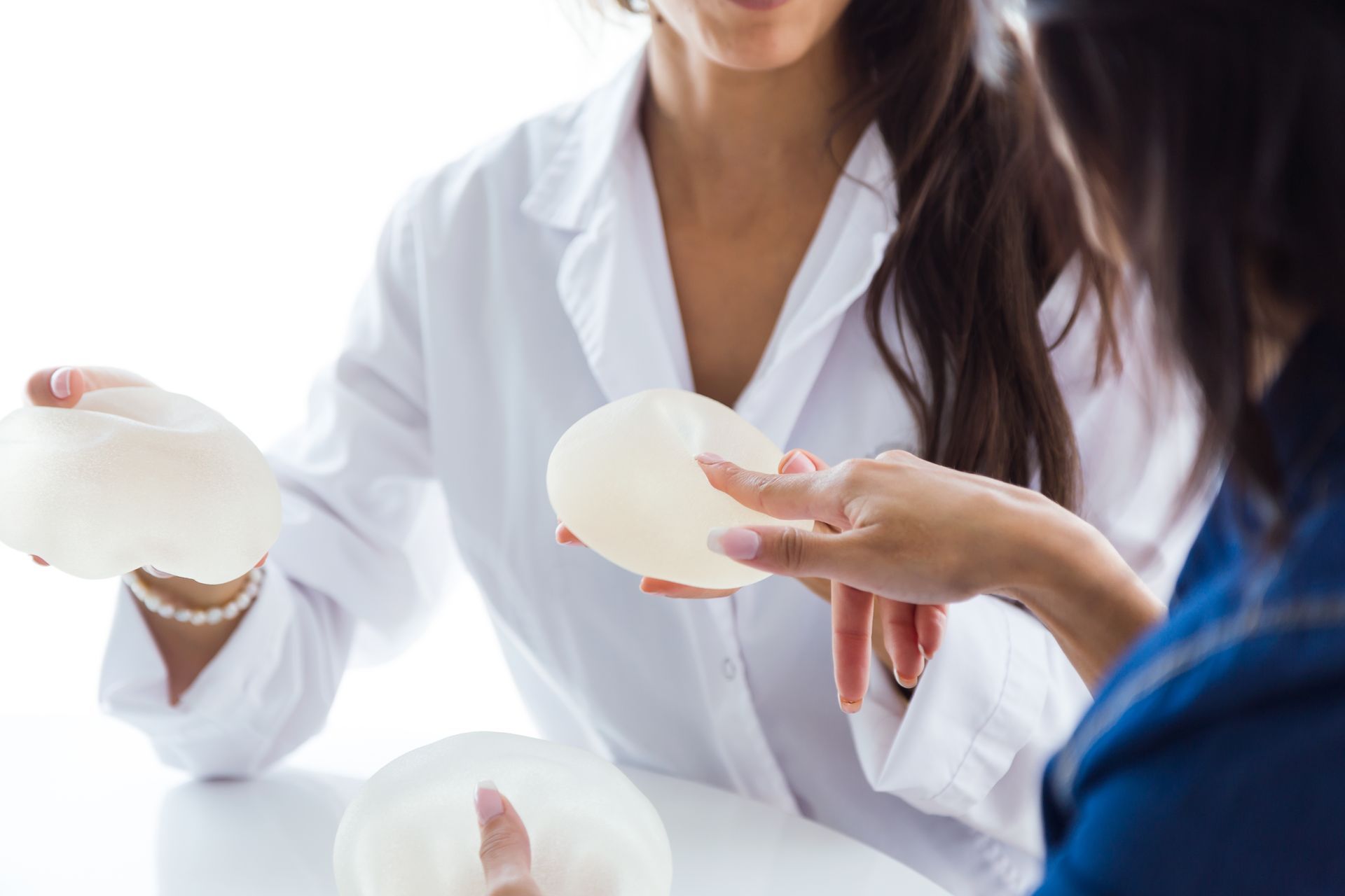 Woman in white coat holding breast implants, consulting with a patient, inside a bright room.