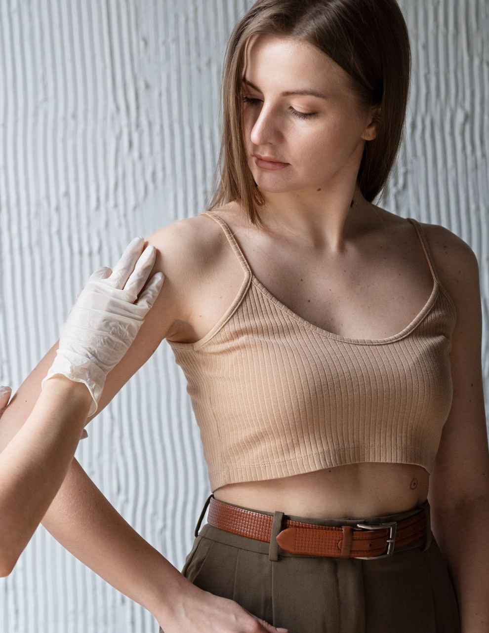 Woman in tan top receiving arm exam; hand with glove touches her shoulder.
