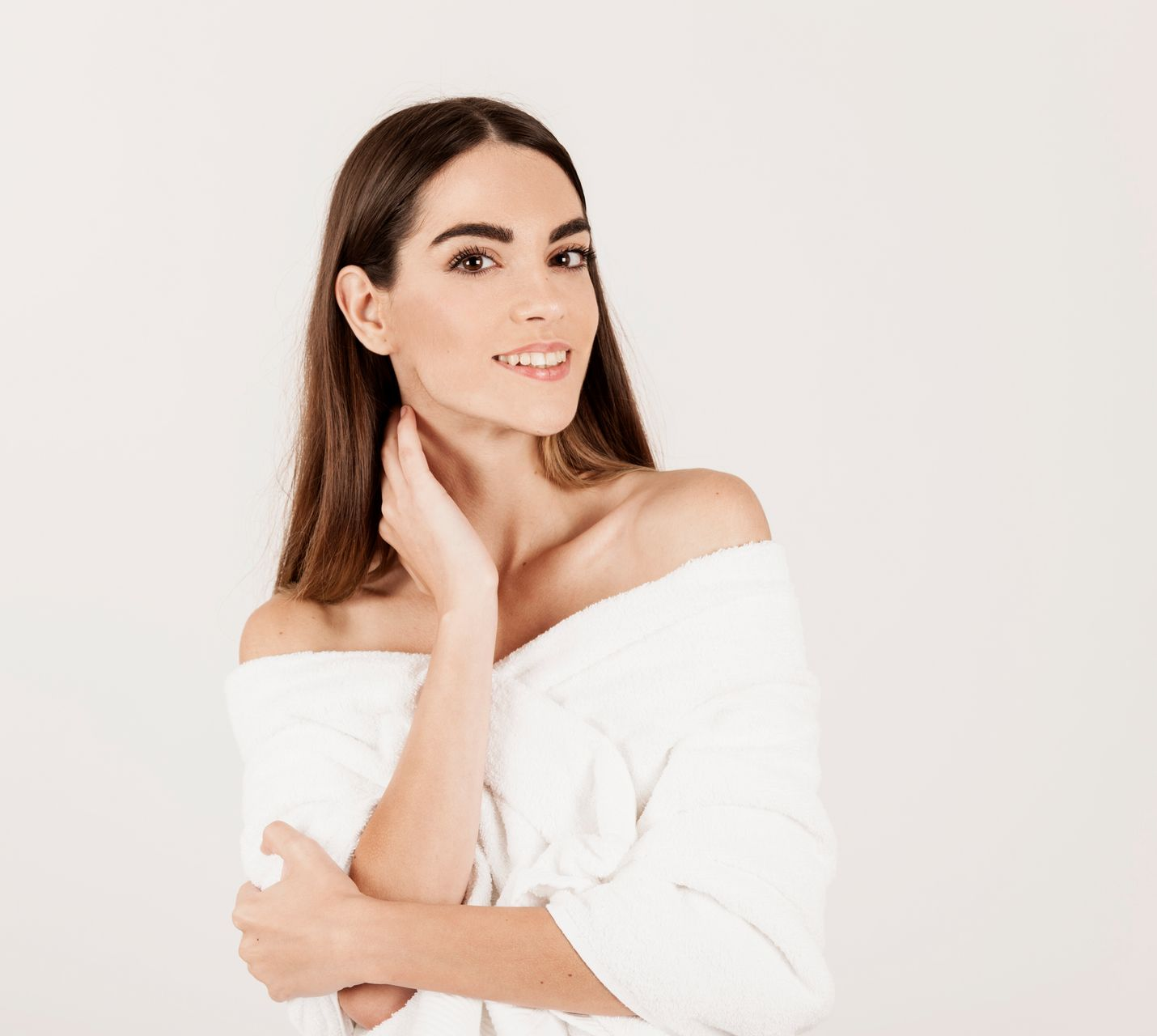 Woman in white robe, smiling, hand on neck, shoulder-baring, against white background.