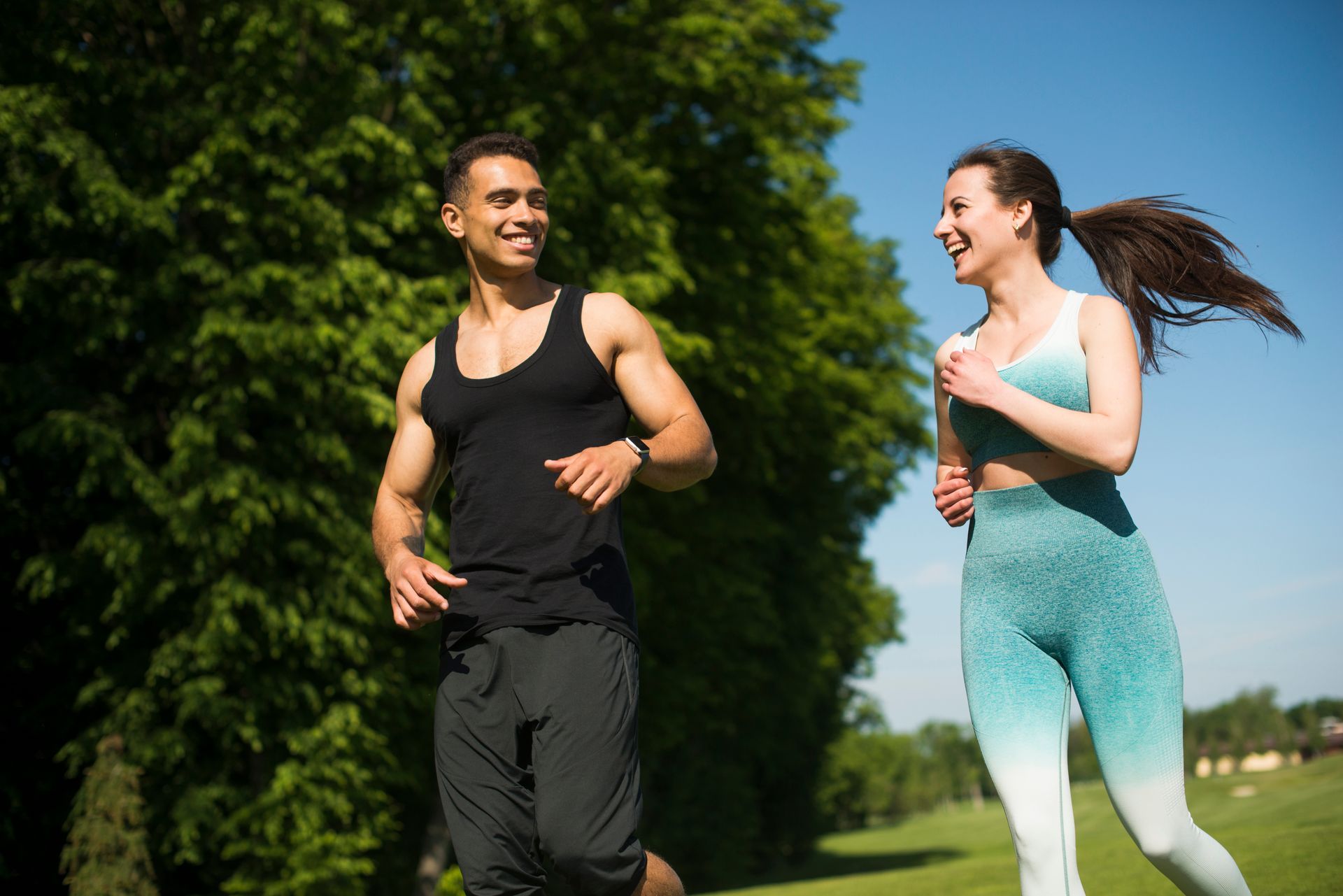 Couple running together in a park, smiling. Man in black, woman in blue activewear, sunny day.