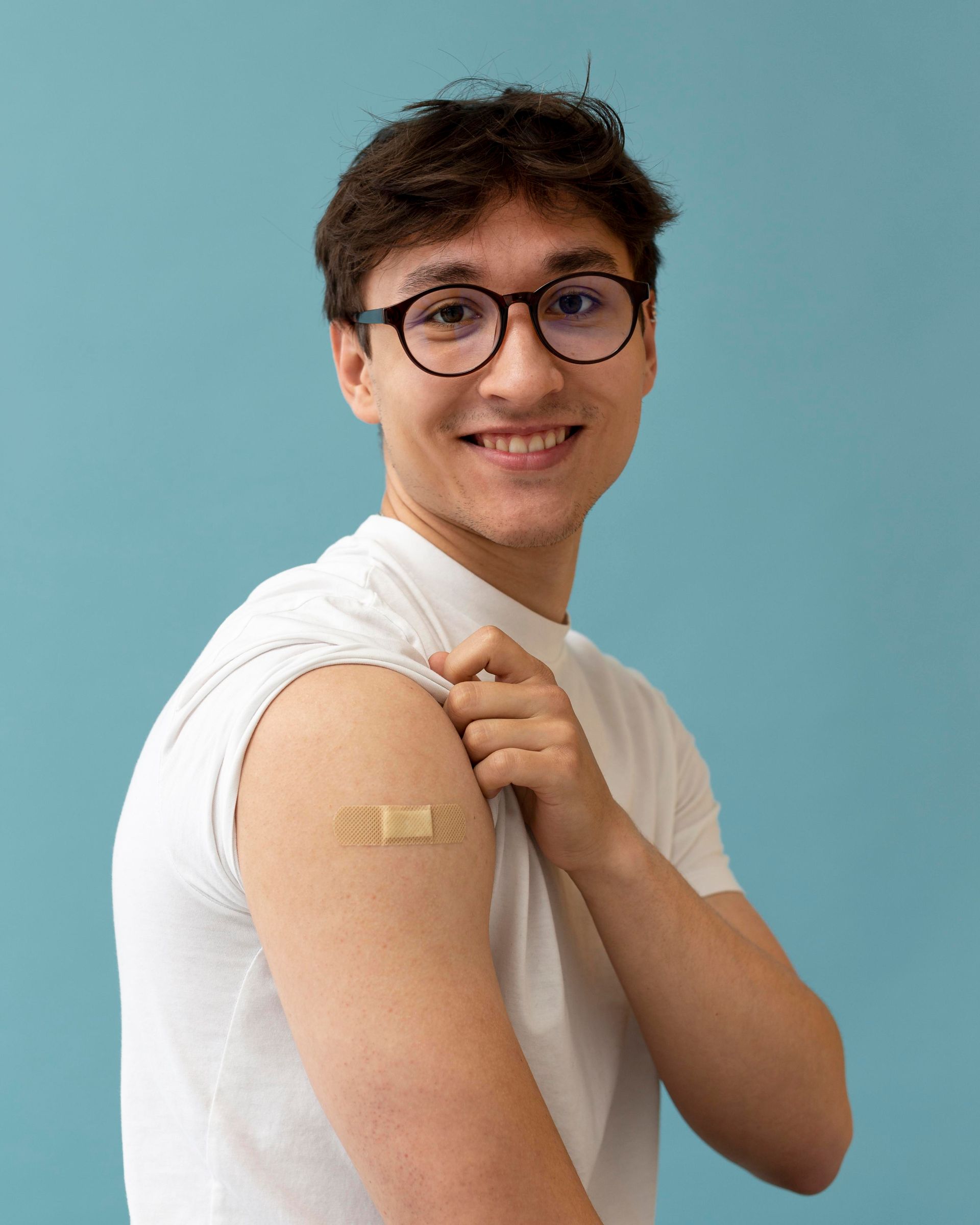 Man with glasses, arm raised, showing a bandage on upper arm, smiling against a blue background.