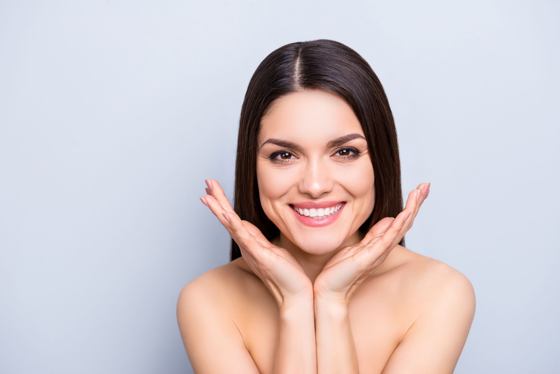 Woman with long dark hair, smiling, hands near her face. Pale background.