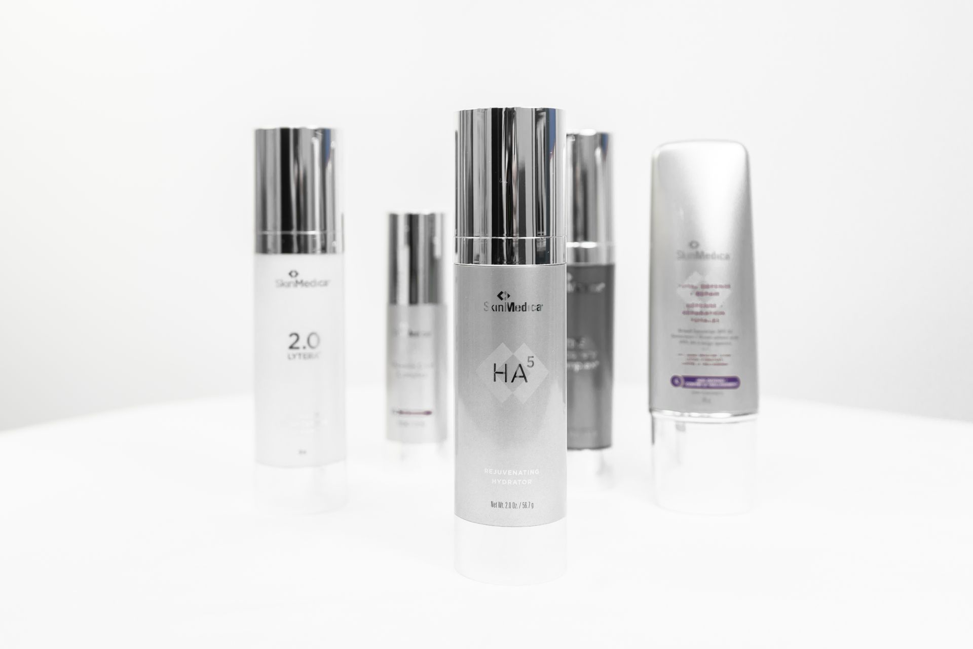 Five silver skincare product bottles on a white surface. Some have the brand name and product type printed on them.