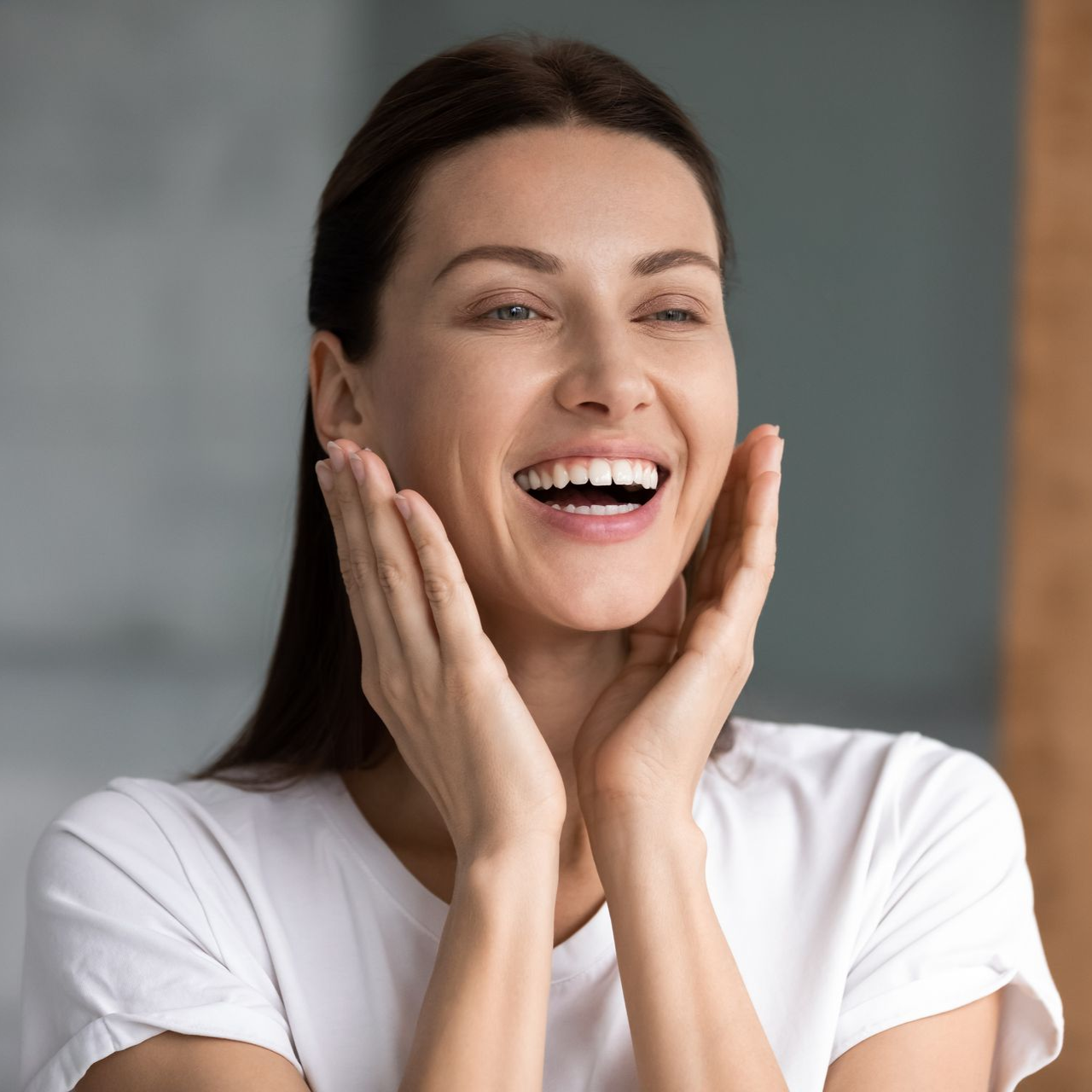Woman with long brown hair, smiling broadly, hands on cheeks, white shirt.