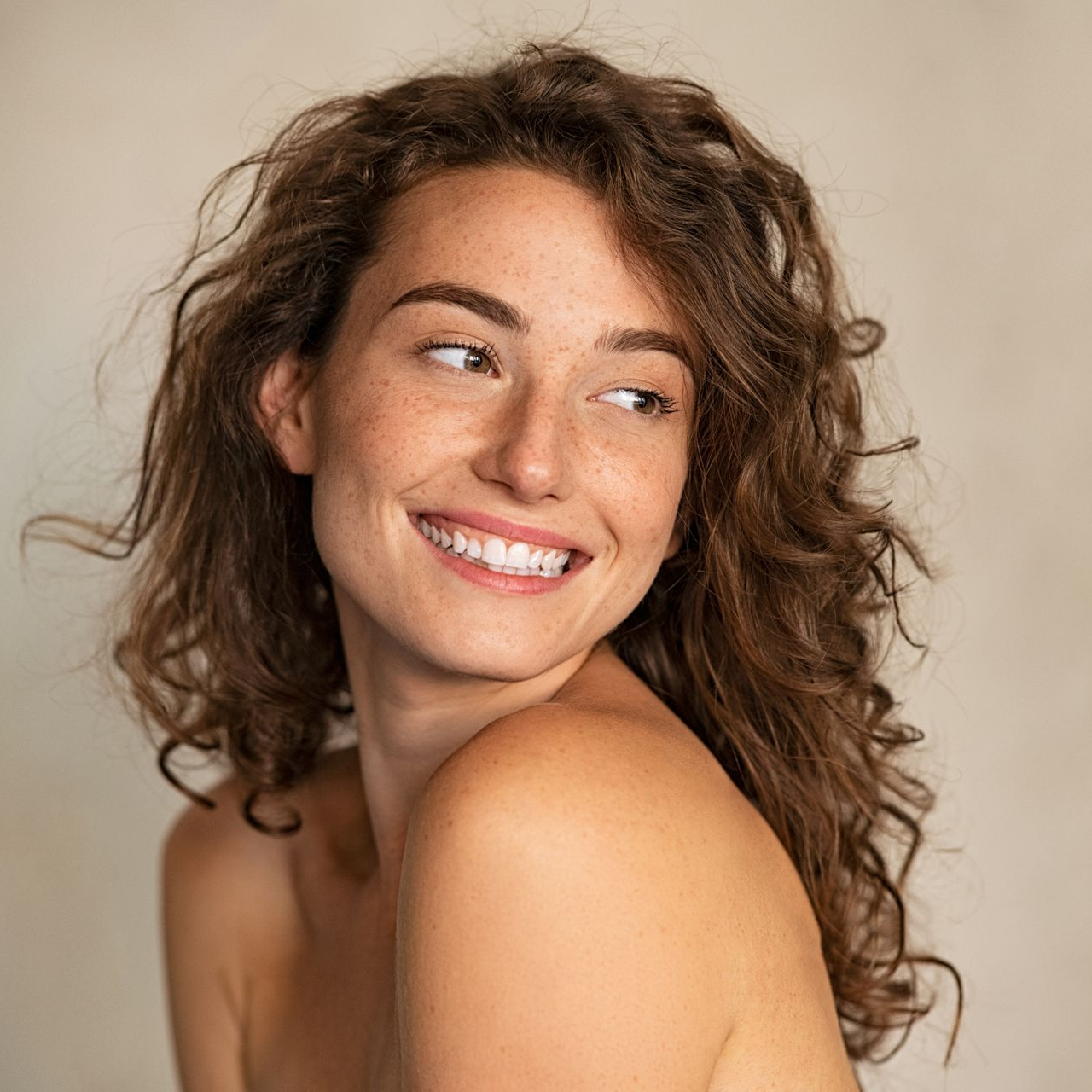Woman with curly brown hair smiles while looking to the side, freckles visible, neutral background.