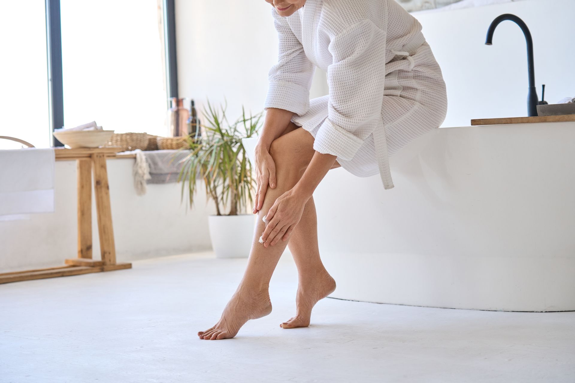 Woman in a bathrobe sitting on a bathtub, applying lotion to her legs in a bright bathroom.