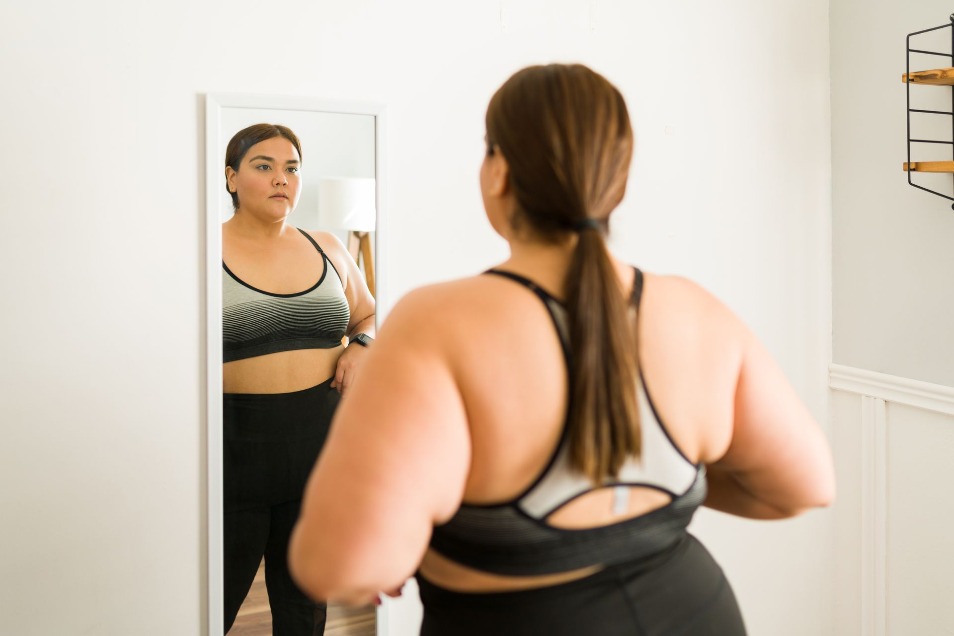 Woman looking in a mirror, wearing a sports bra and leggings. Interior shot.