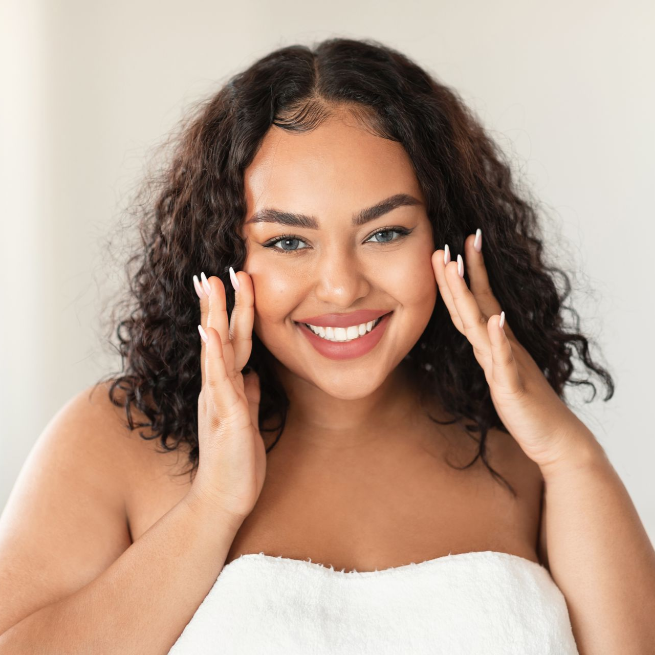 Woman with curly hair smiling, touching her face wrapped in a towel.