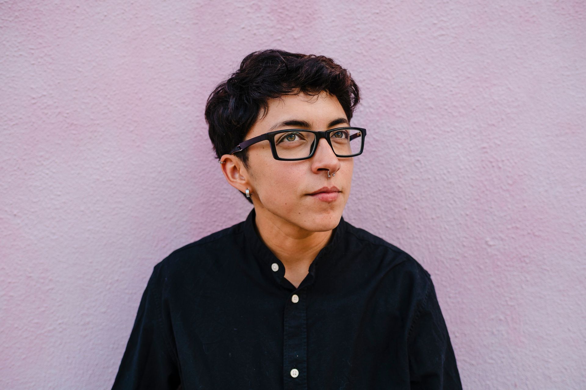 Person with dark hair and glasses looking off-camera, wearing a black shirt, standing against a pink wall.