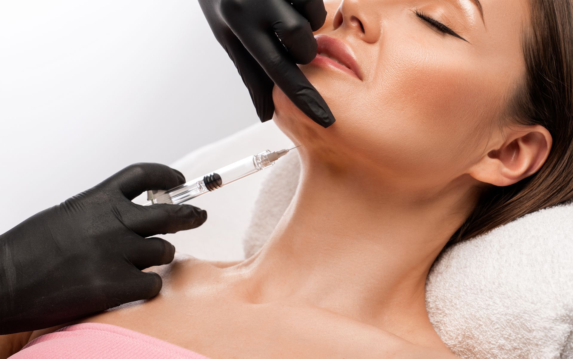 Woman receiving a cosmetic injection in the chin. Black-gloved hands, white background.