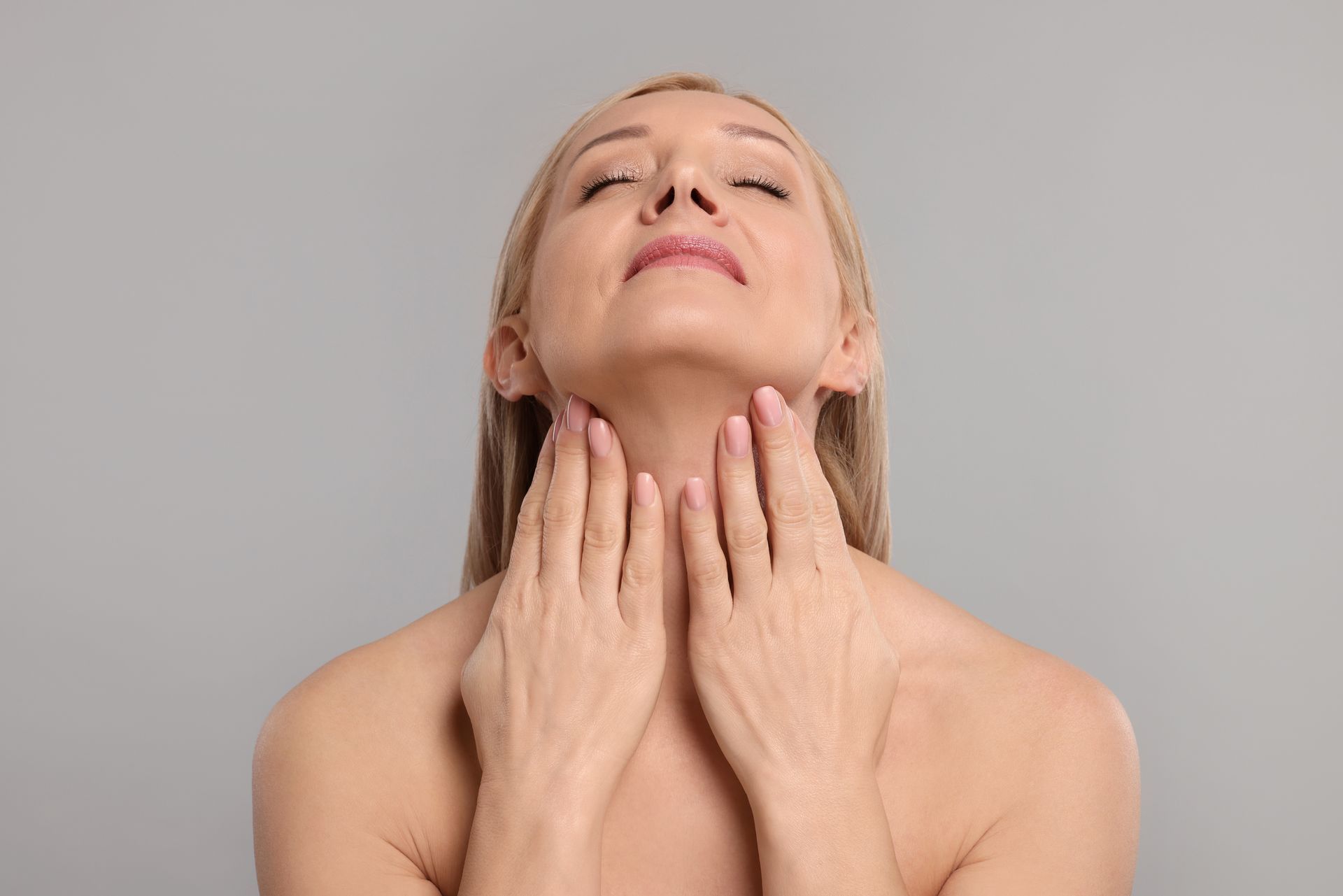 Woman touching her neck, eyes closed, with visible wrinkles on her neck and hands. Gray background.