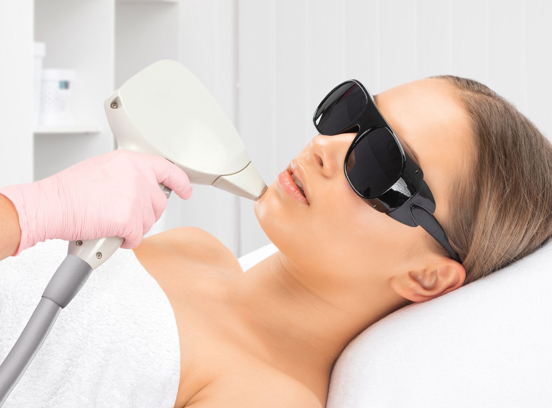 Woman receiving laser hair removal treatment on her chin, wearing protective eyewear, in a clinical setting.