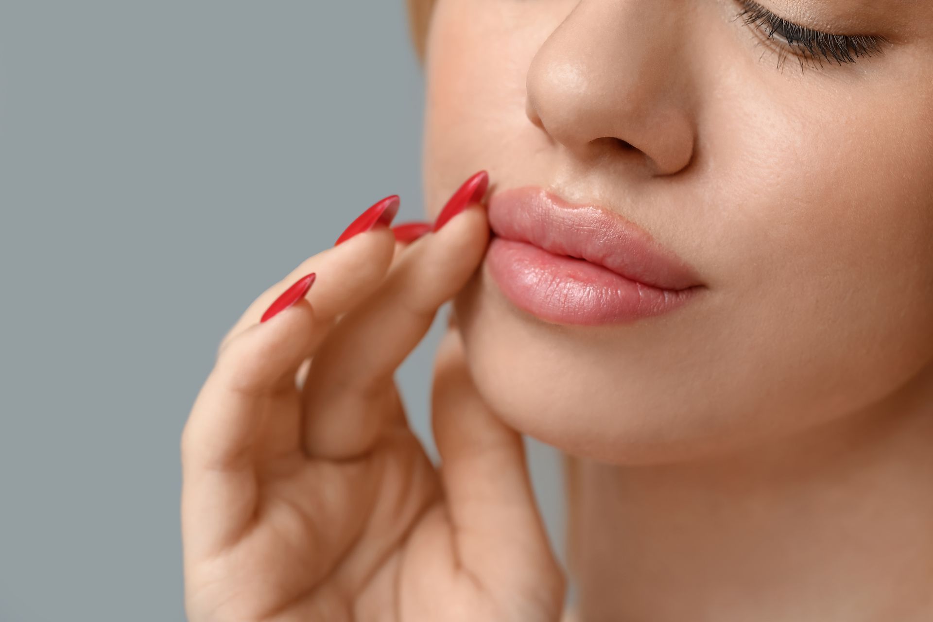 Woman touching her glossy lips with red fingernails; close-up with gray background.