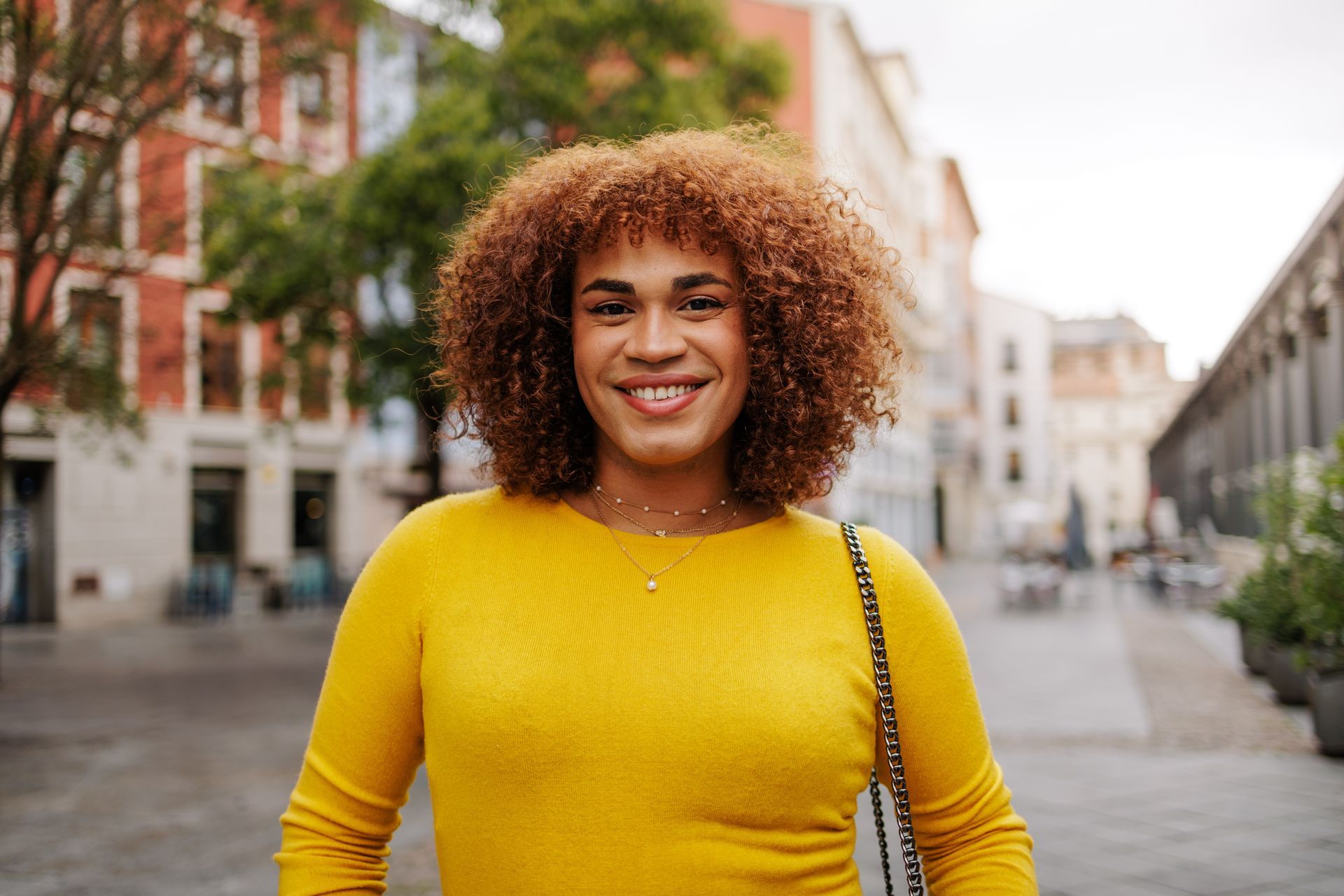 Person smiling outdoors, wearing yellow sweater, curly hair. City background.