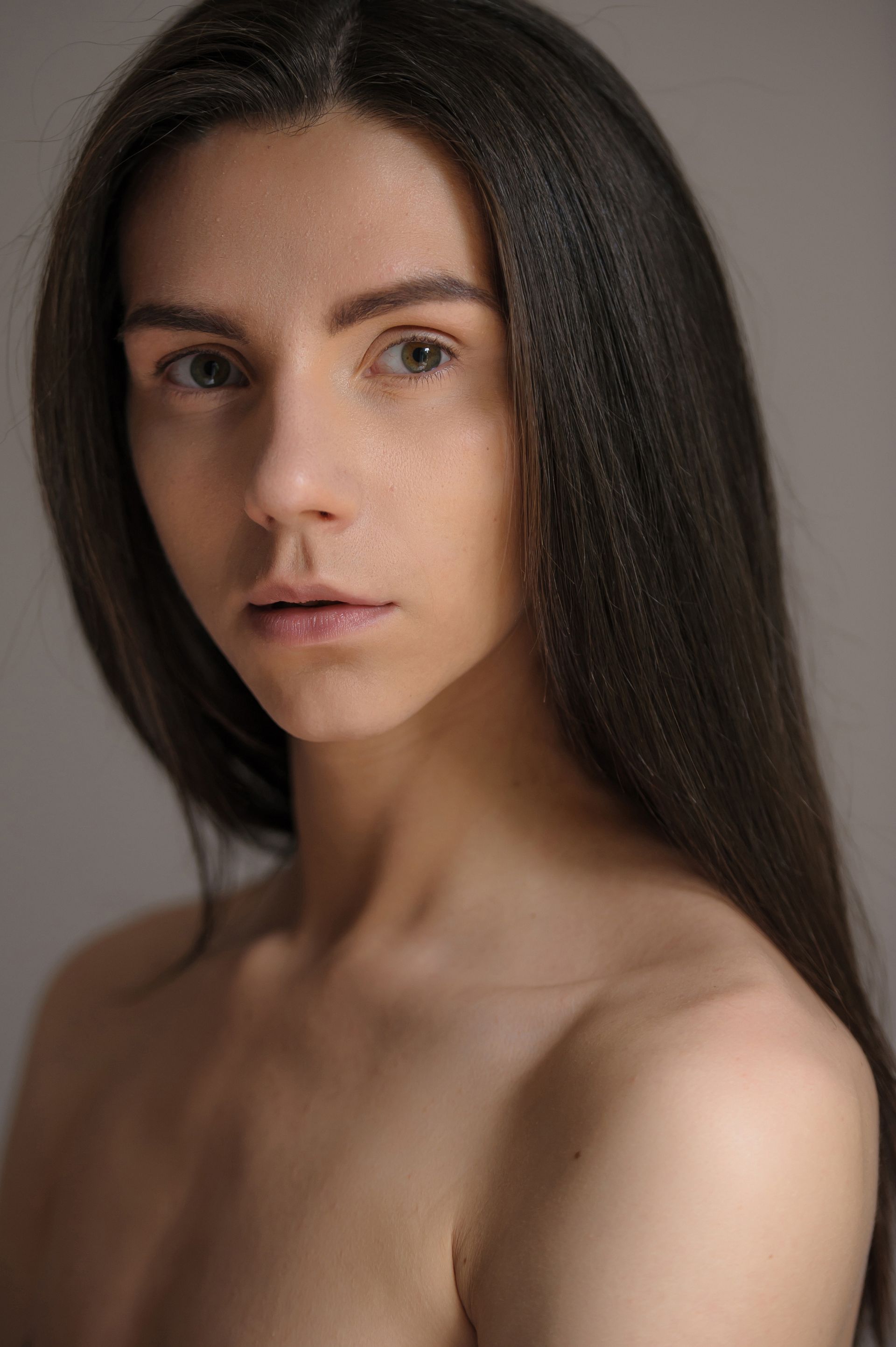 Person with long dark hair looking directly at the viewer, bare shoulders, neutral background.