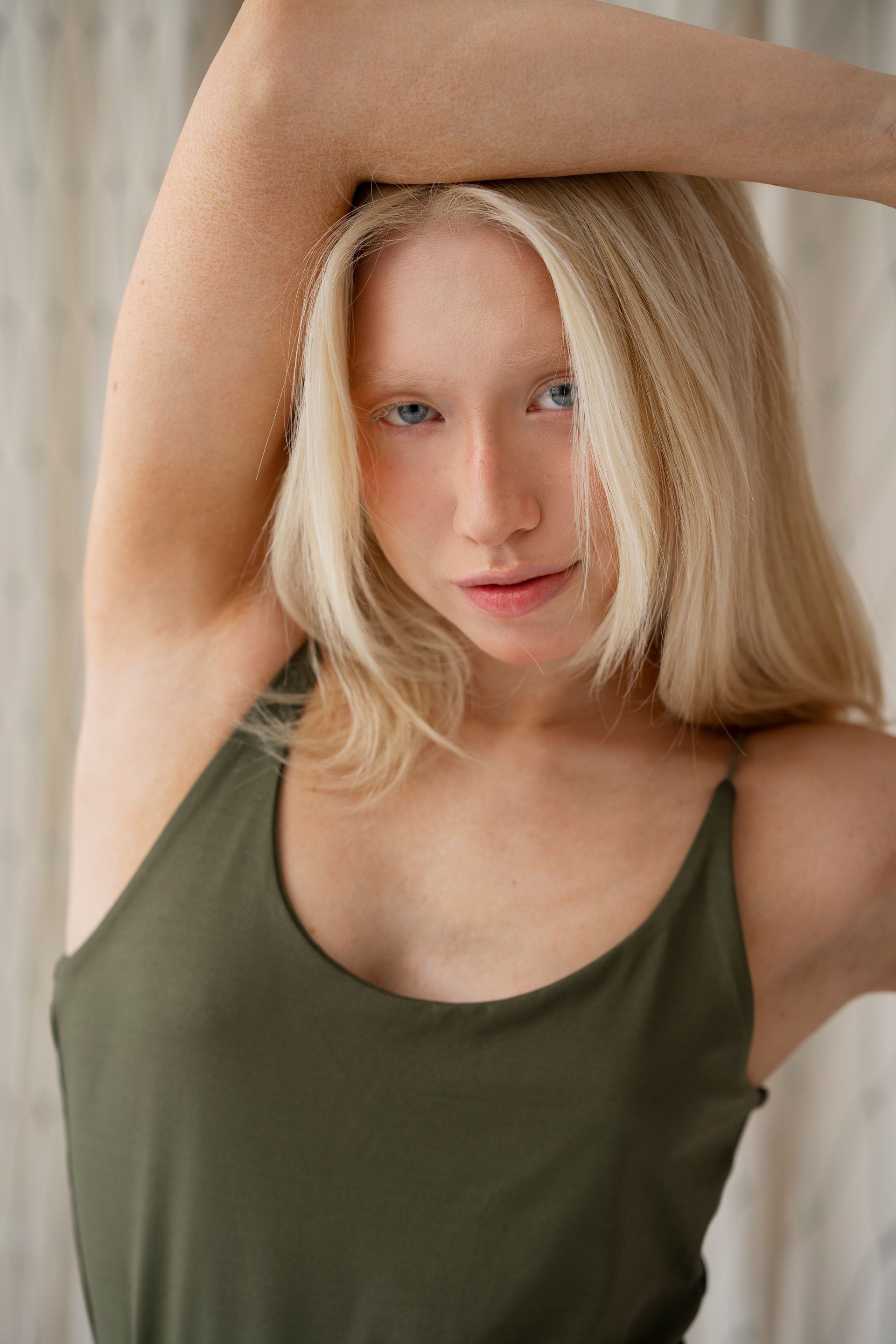 Blonde person with blue eyes in a green tank top, arms raised, facing forward.