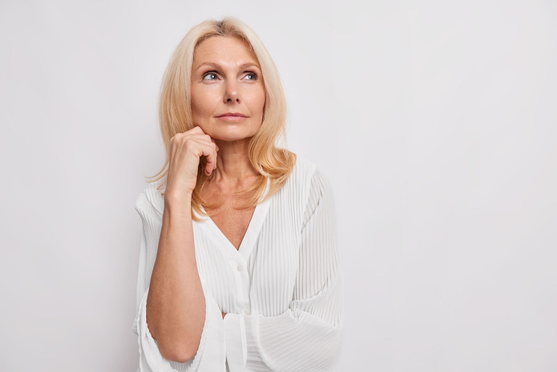 Woman with blonde hair looks upward, hand on chin, wearing a white blouse, against a white background.