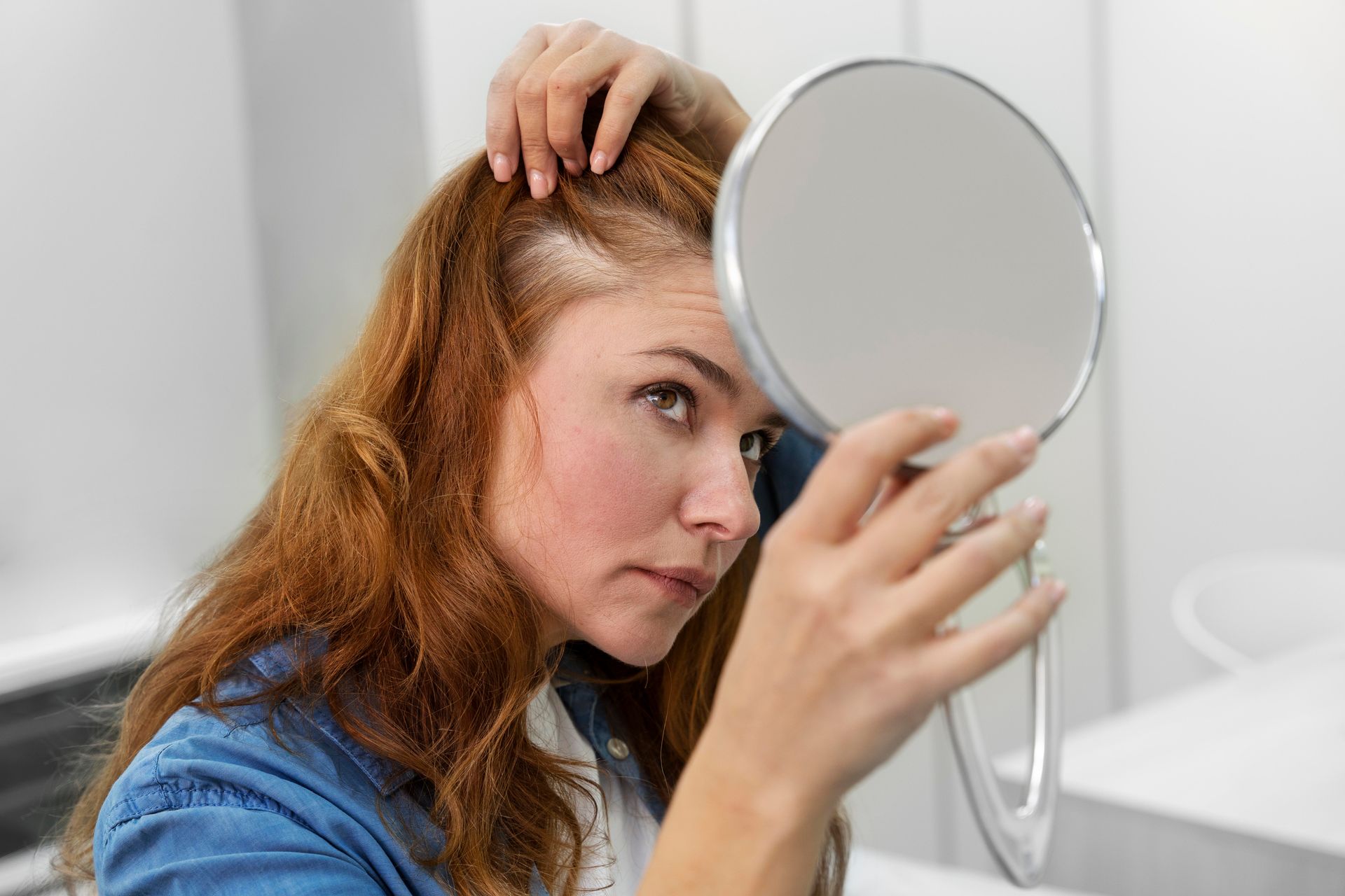 Woman examining thinning hair with a hand mirror. Red hair, denim jacket, white wall background.