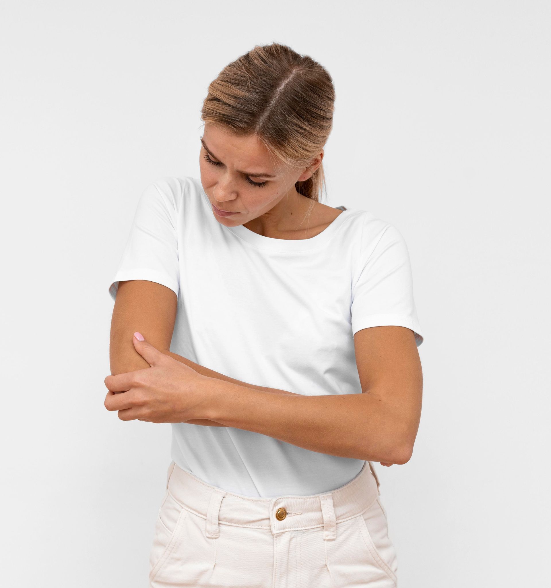 Woman in white shirt and tan pants holding her elbow, looking down with a concerned expression.