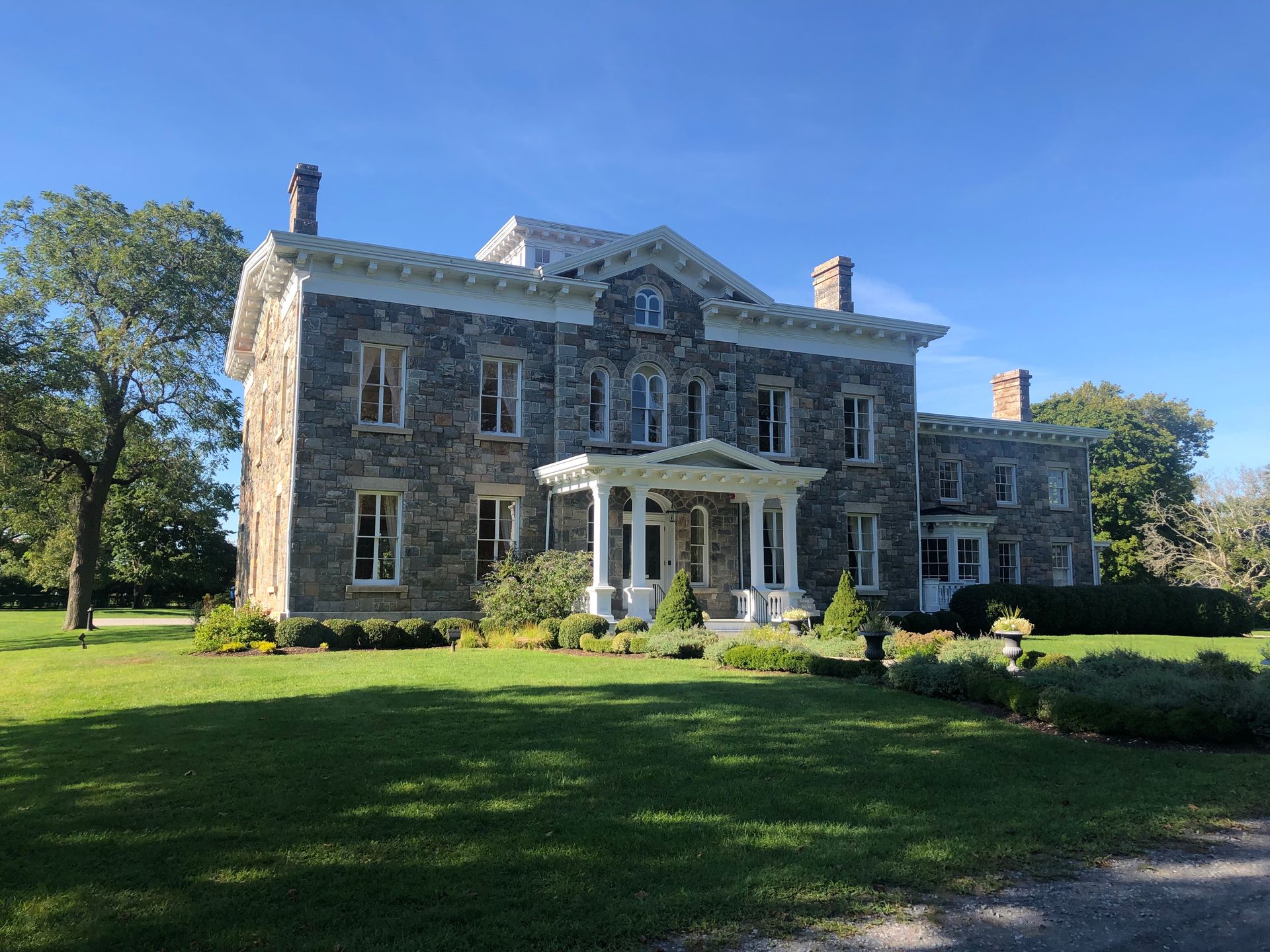 A large stone house with a white porch is sitting on top of a lush green field.