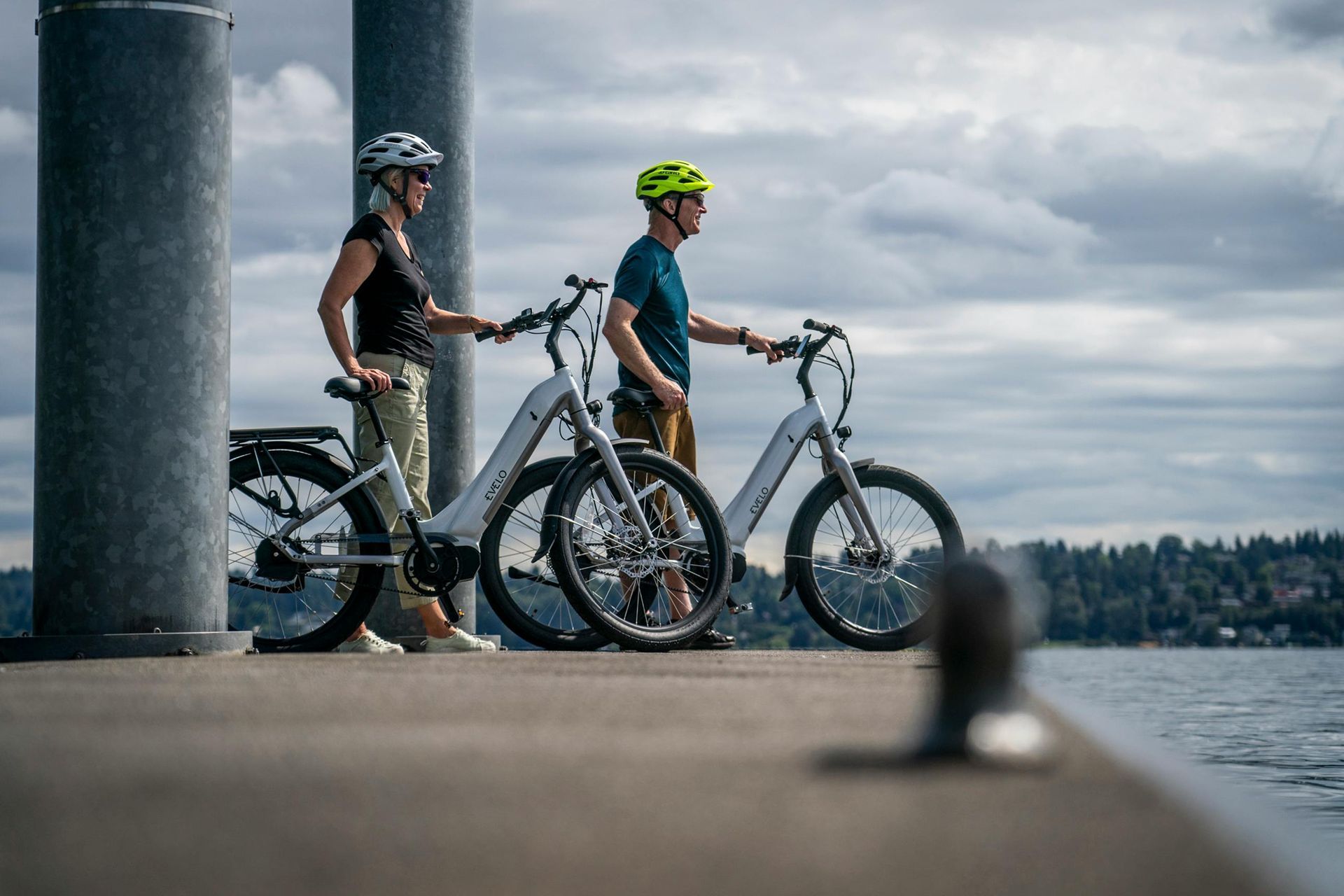 A man and a woman are standing next to their electric bikes on a dock.