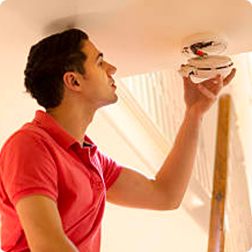 A man in a red shirt is installing a smoke detector on the ceiling.