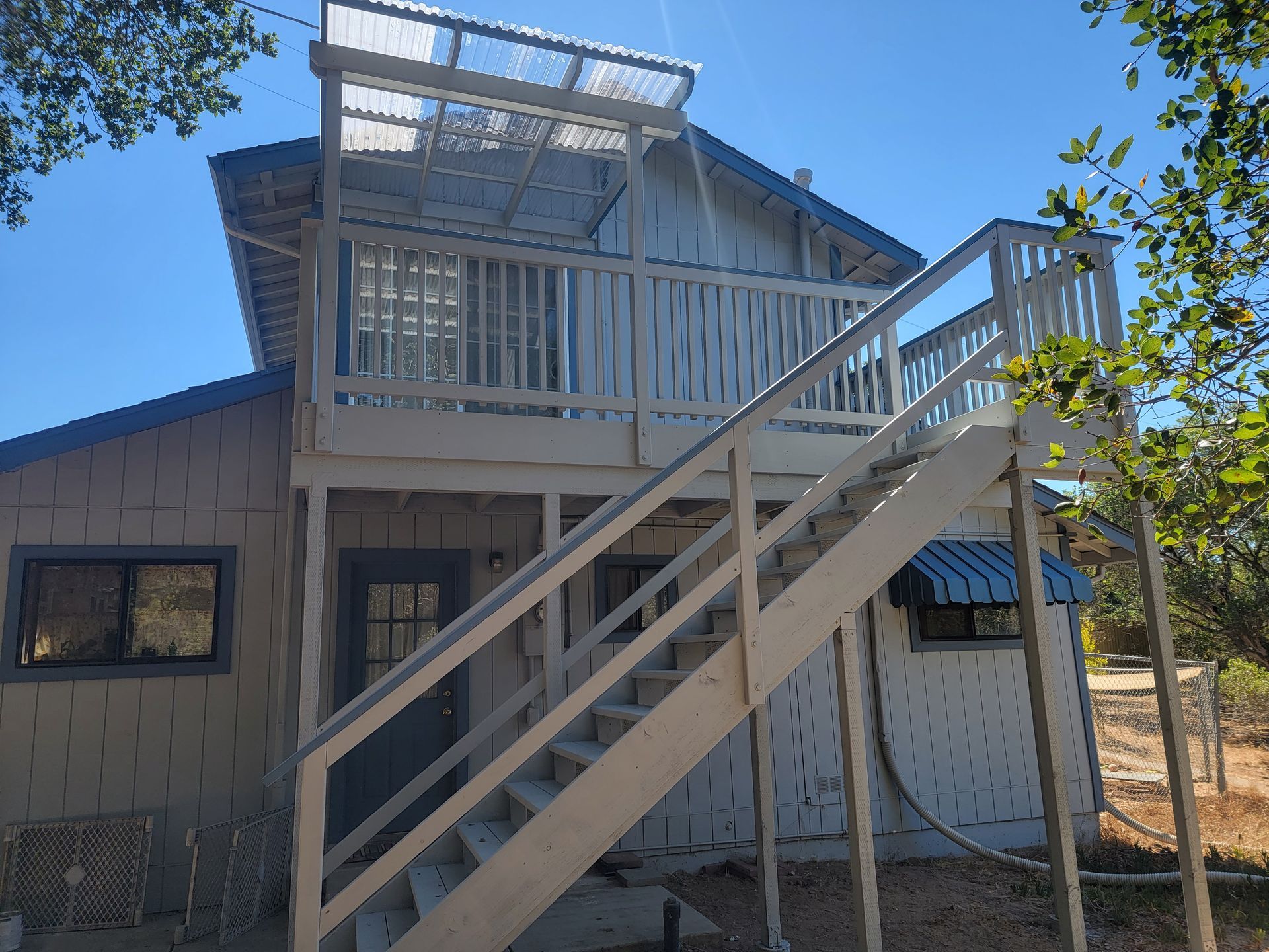 Two-story wooden house with exterior stairs leading to a deck; blue sky overhead.