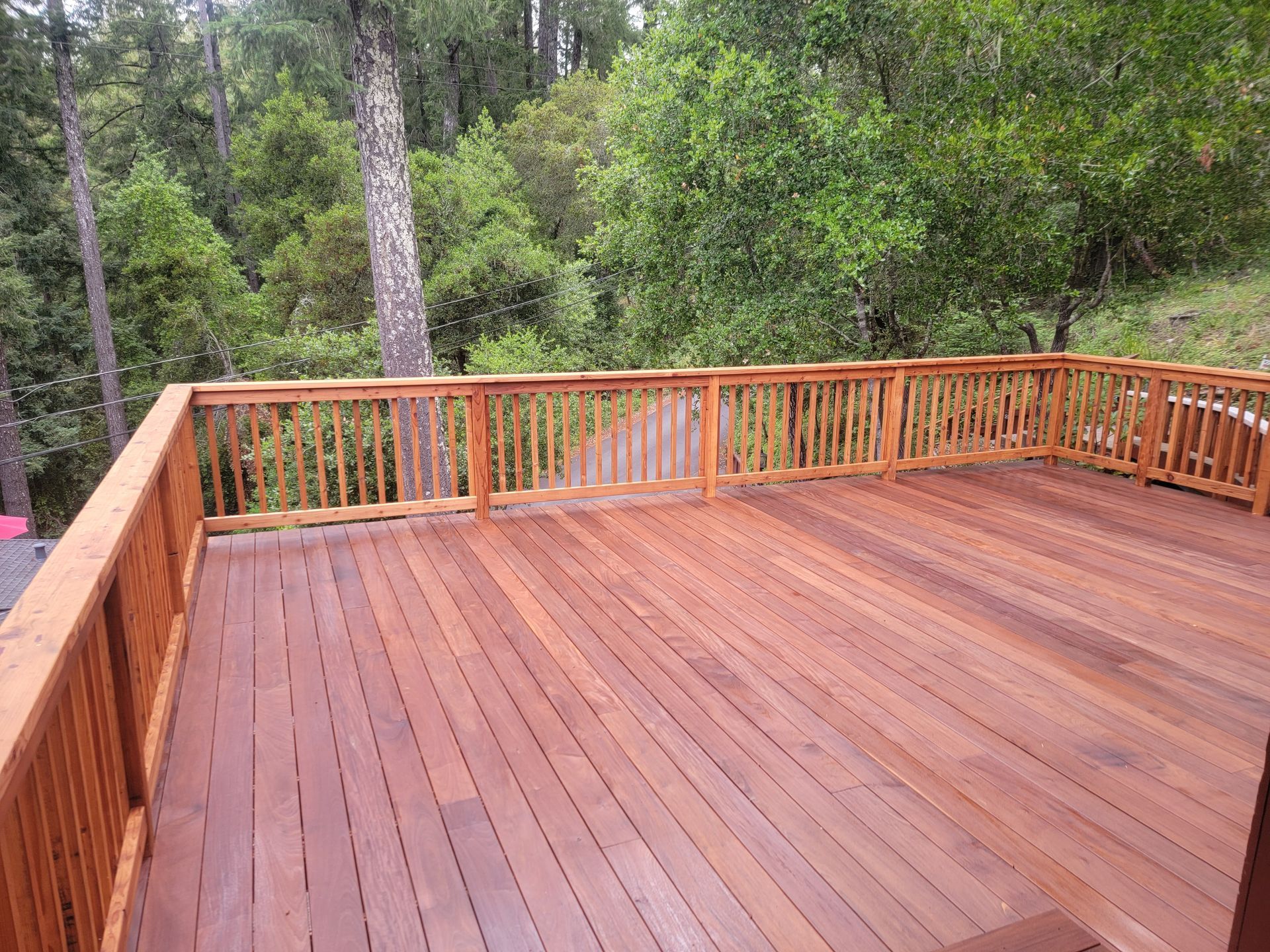 Wooden deck with railing overlooking a wooded area. Brown wood and green trees.