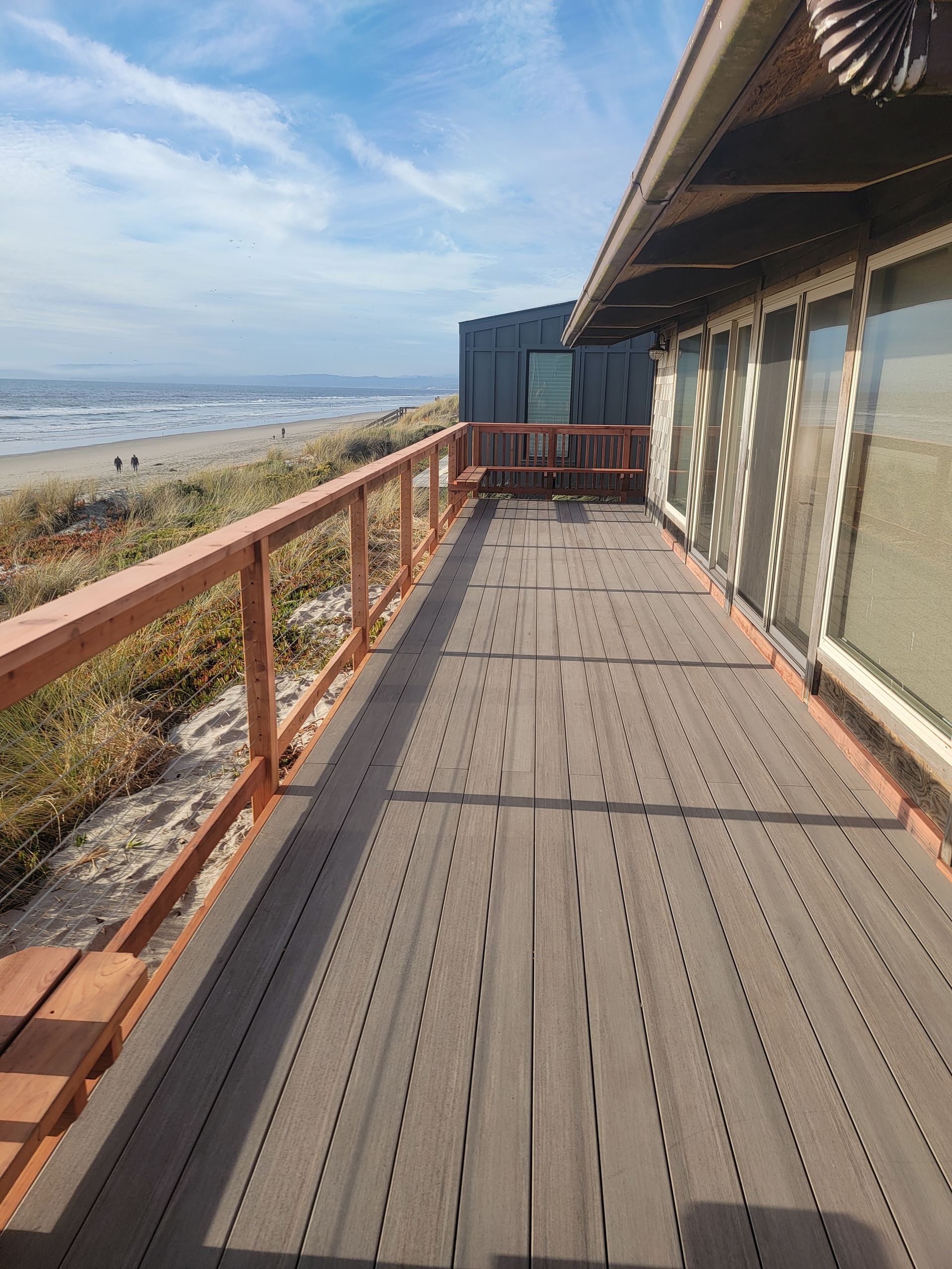 Wooden deck extends toward the ocean beside a building. Brown railing, clear sky, sandy beach.