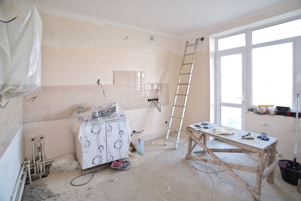 Kitchen renovation in progress: walls prepped, ladder, tools, covered appliances, and bright window.