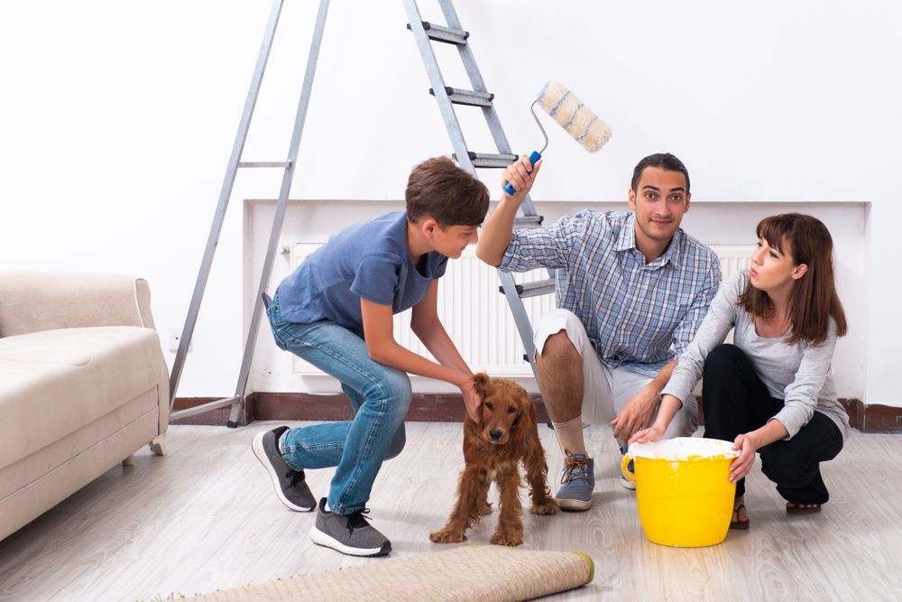 Family painting a wall with a dog. Boy pets dog, father uses roller, mother holds paint bucket.
