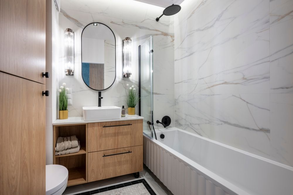 Modern bathroom with wood vanity, oval mirror, white marble tile, and bathtub.