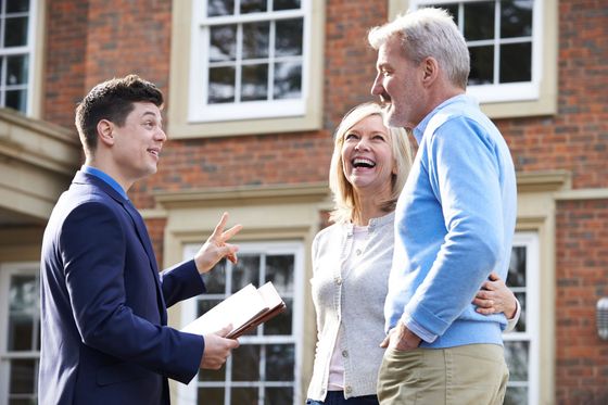 Real estate agent talking to a smiling couple in front of a brick house.