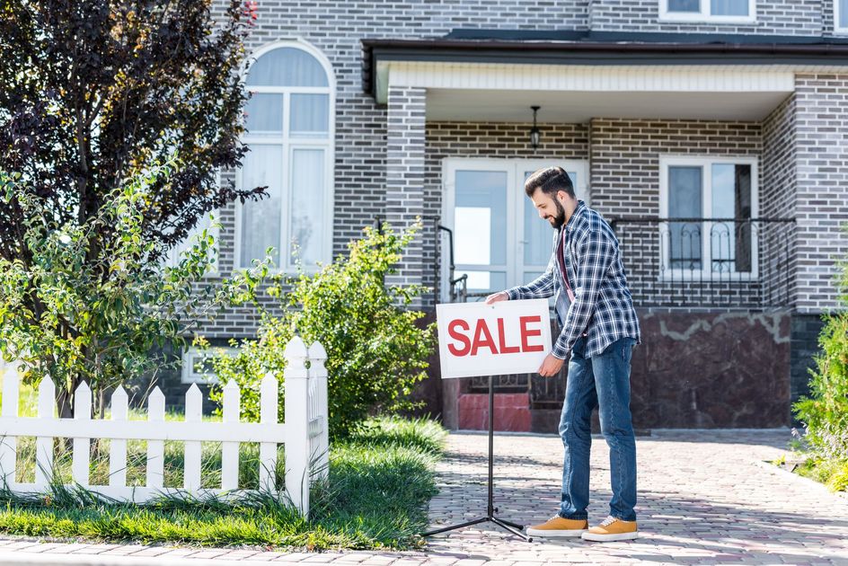 Man placing a SALE sign in front of a brick house.