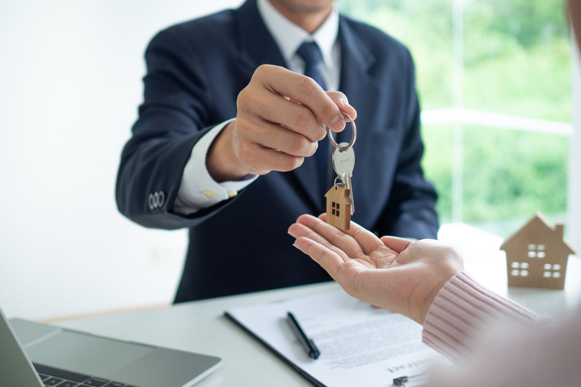Person in suit handing keys to another person over a desk. A h ouse model and paperwork are visible.