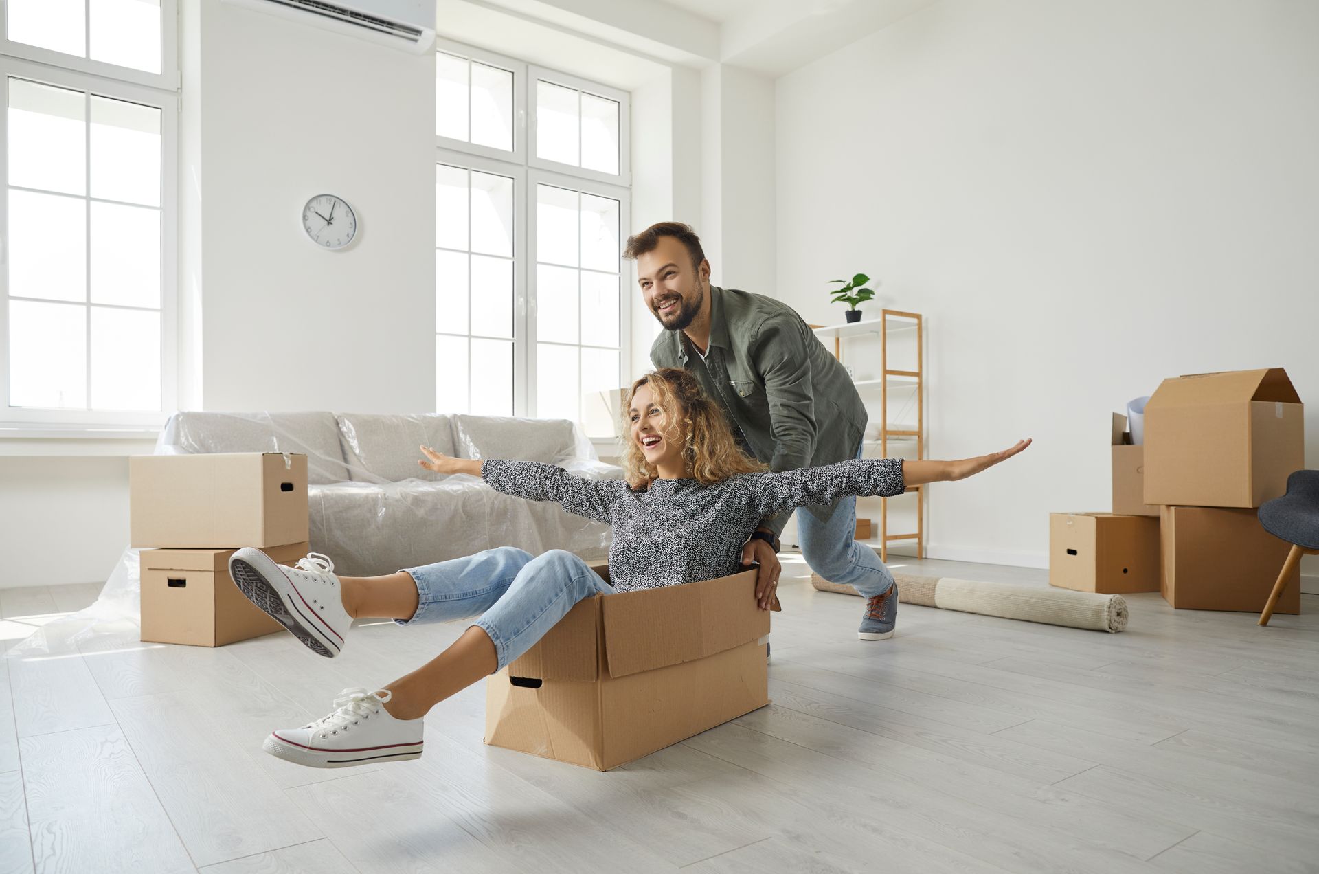 Man pushing woman in a cardboard box, arms outstretched, in a new home with boxes and a sofa.