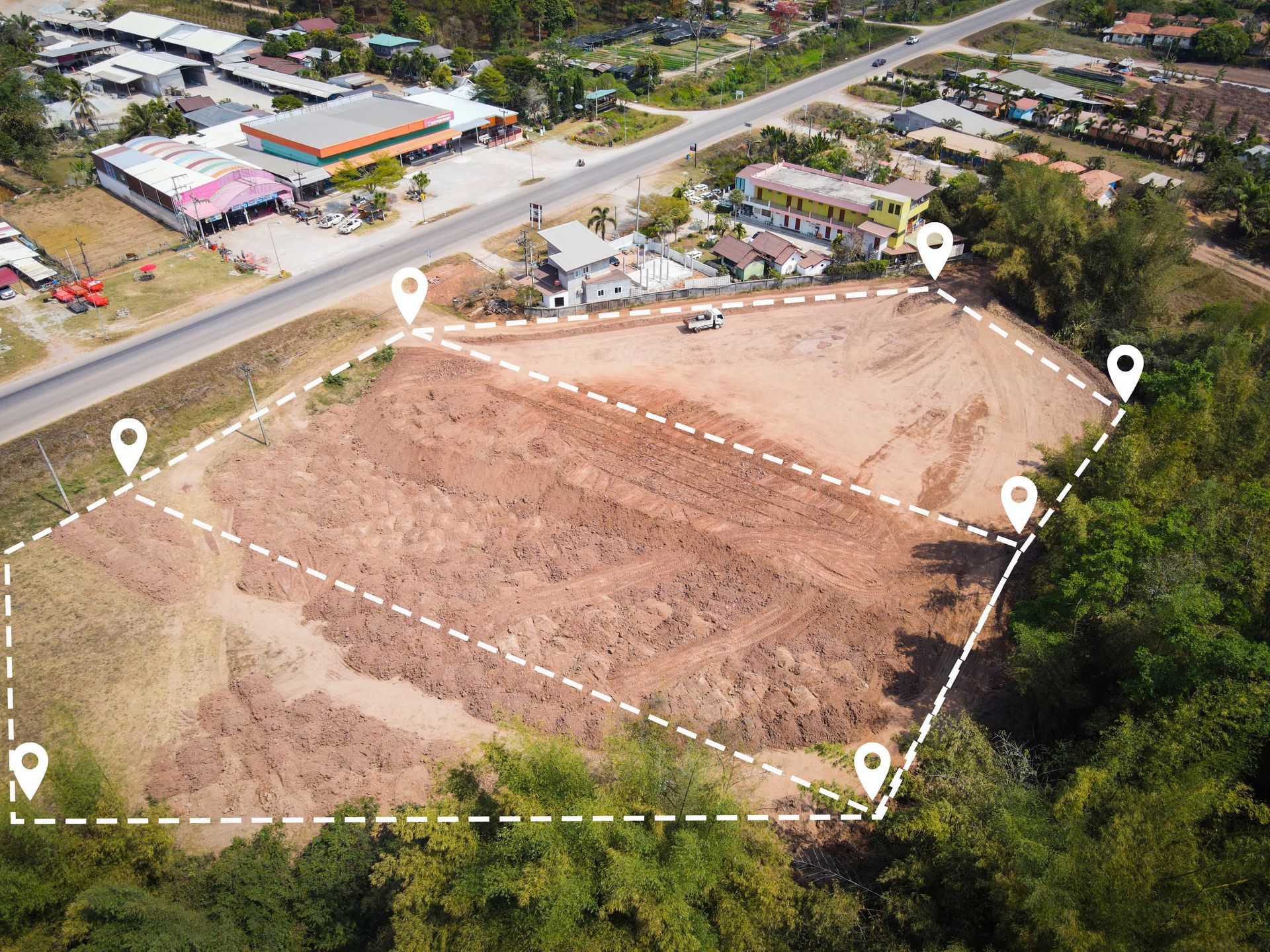 Aerial view of a cleared plot of land with white dotted boundary lines and pin markers; a road and buildings are in the background.