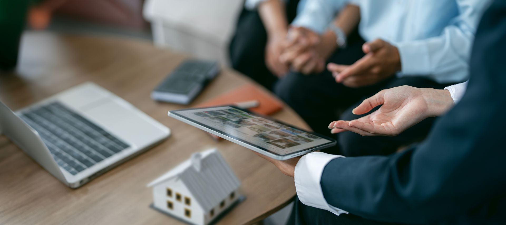 Real estate agent pointing at a tablet screen while discussing with clients; small house model and laptop on the table.