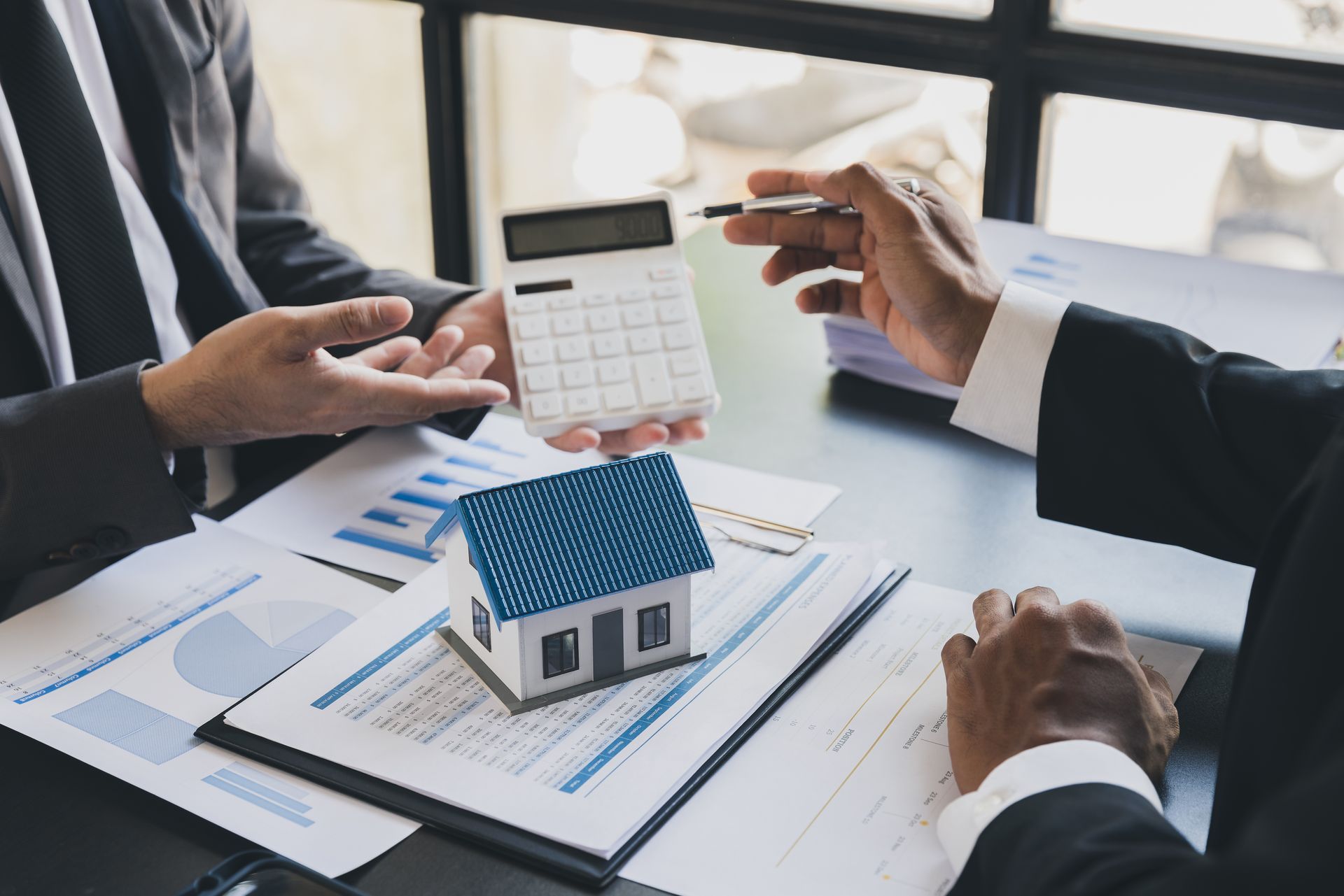 Two people in suits review paperwork with a model house and calculator on a desk.