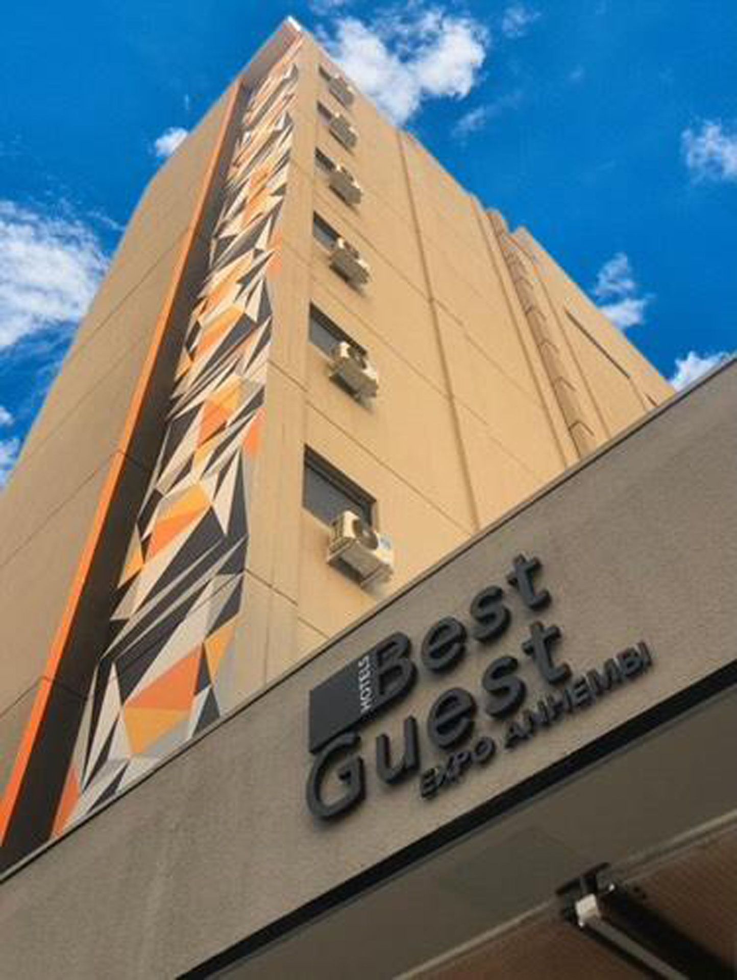 Vista em ângulo baixo do hotel Best Guest Expo Anhembi, um prédio alto bege com design geométrico e céu azul.