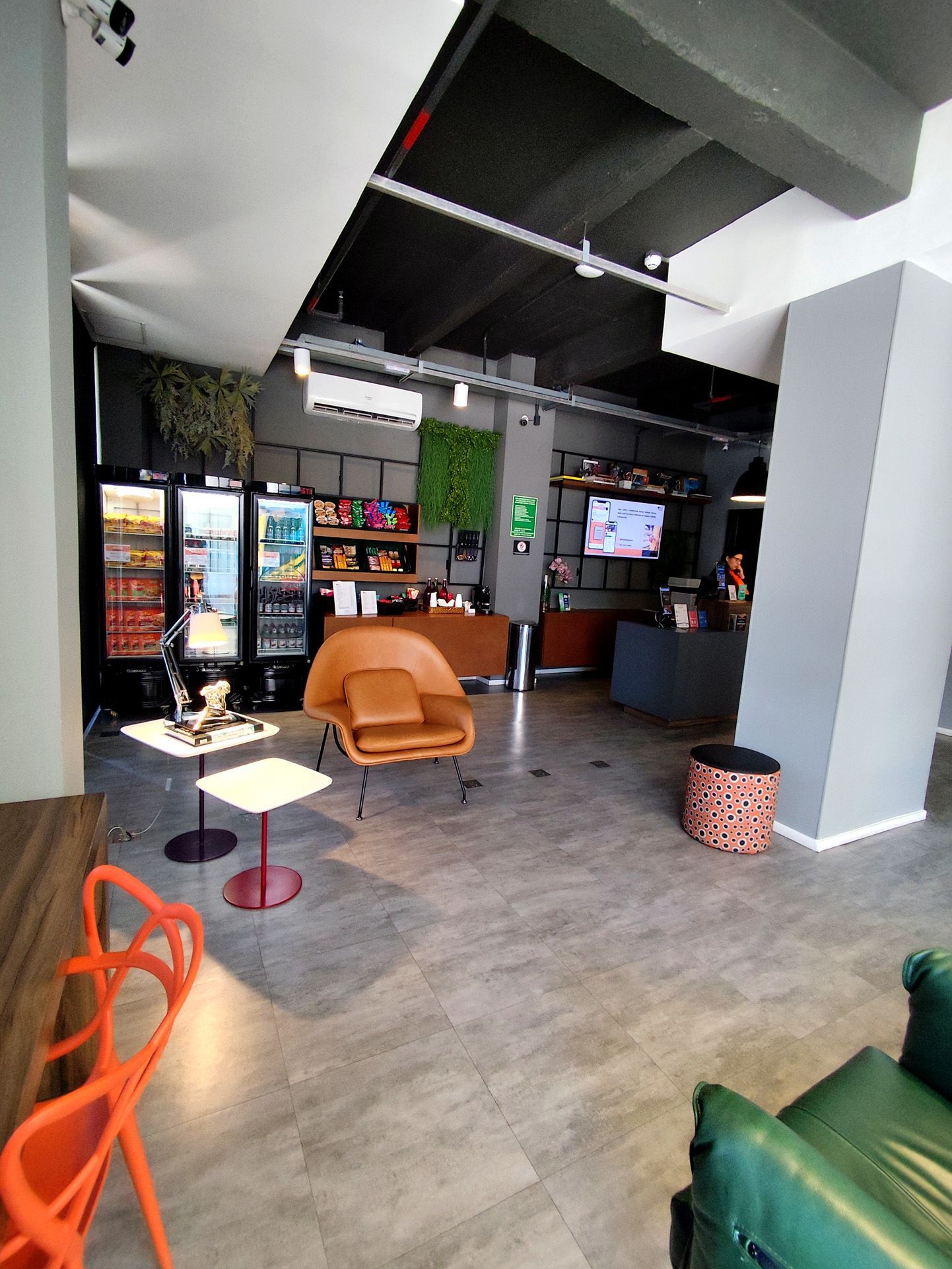 Área de estar com assentos, expositores de lanches e TV. Apresenta tons de laranja, verde e cinza.