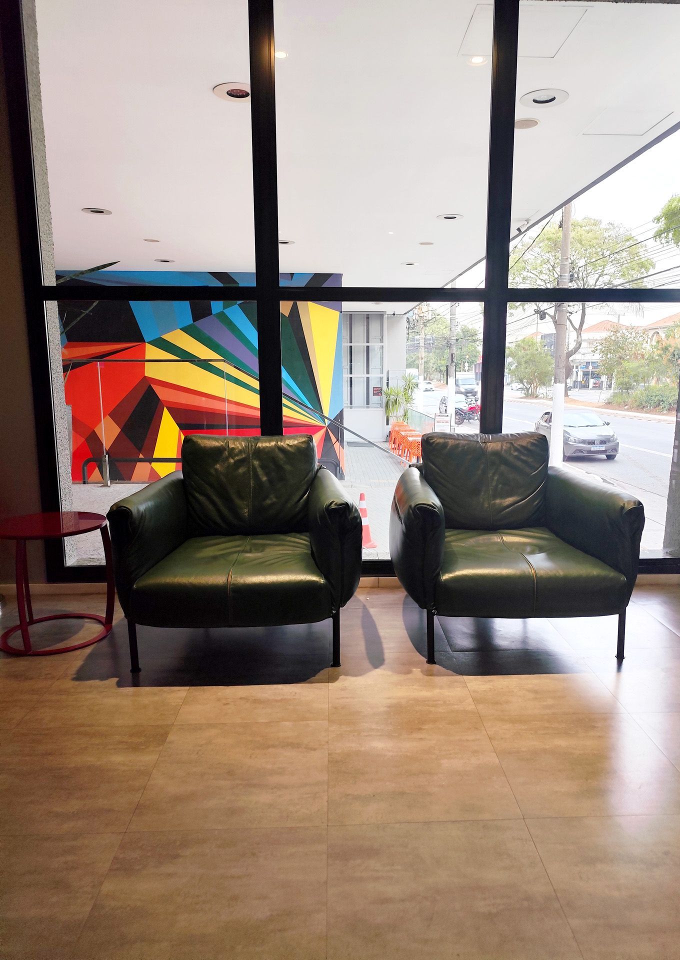 Duas poltronas verdes ficam perto de uma grande janela, com um mural abstrato colorido visível através do vidro.