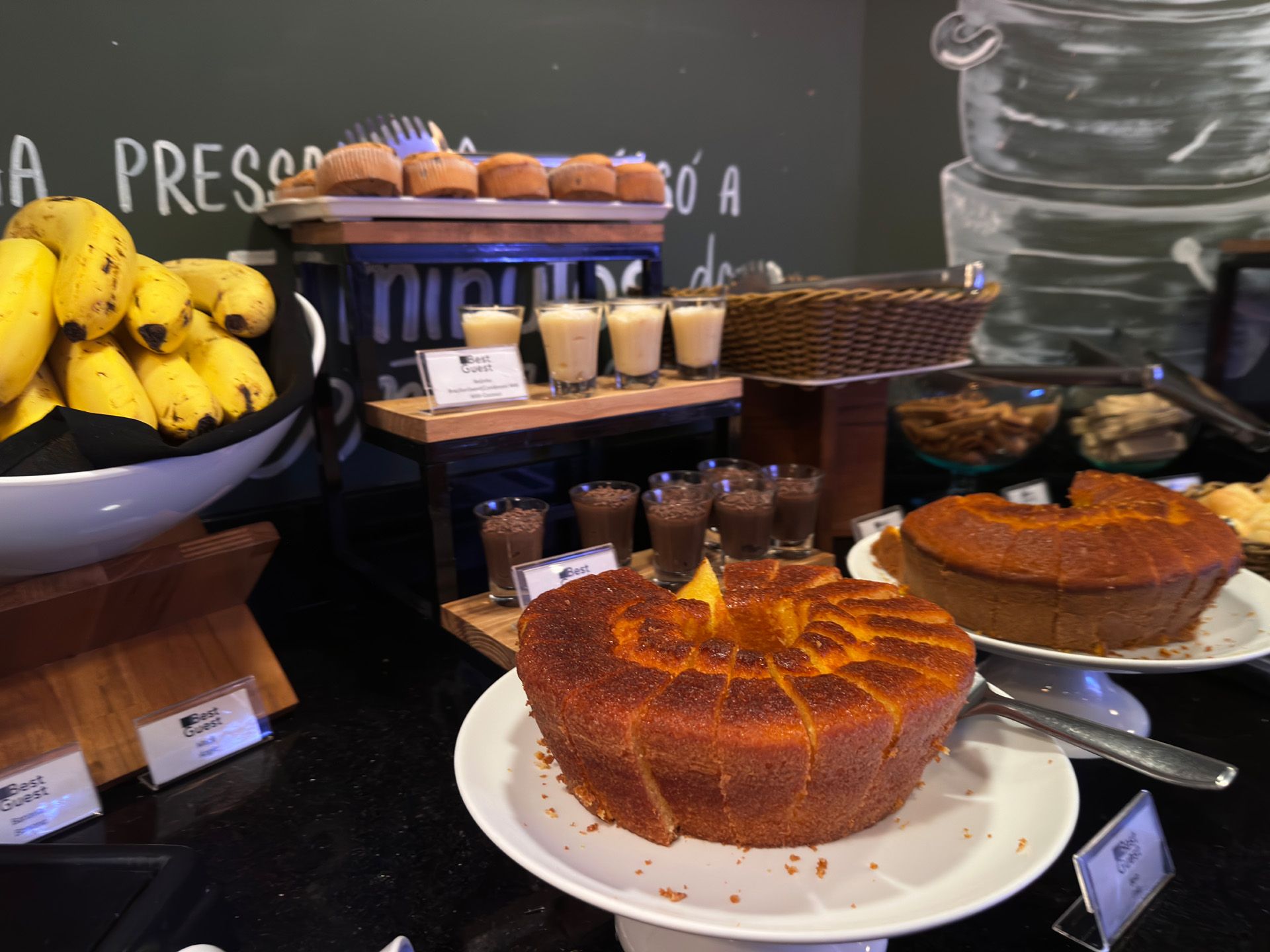 Buffet de café da manhã com bolos, bananas e bebidas pequenas, disposto sobre uma superfície preta.
