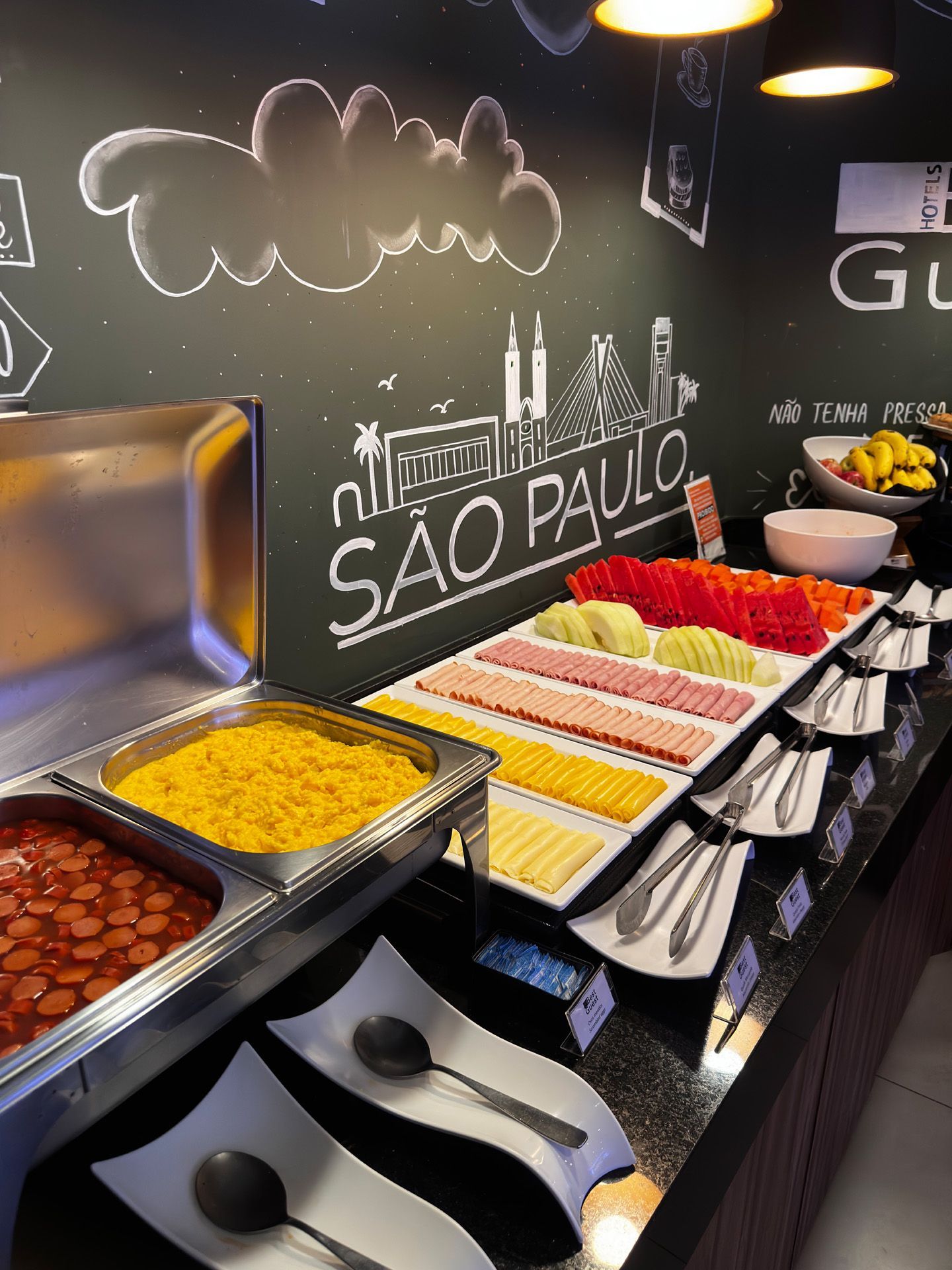 Buffet de café da manhã com diversas opções de comida em um balcão, fundo preto com desenho de São Paulo.