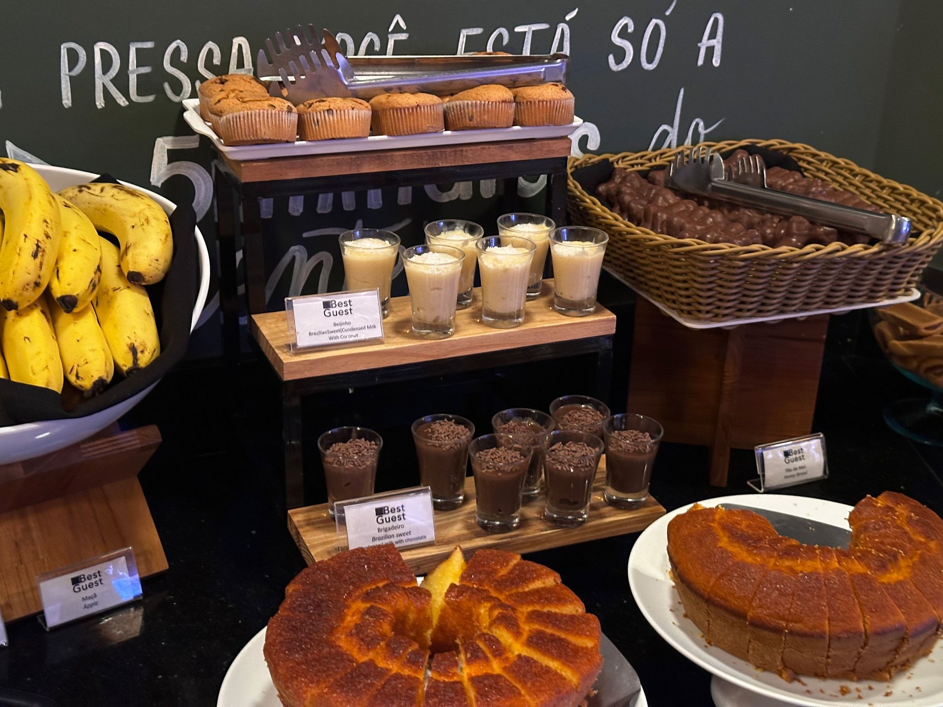 Buffet de café da manhã: bananas, muffins, bolo, pequenas porções de sobremesa e uma cesta de pães.